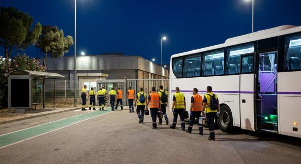 Relevo nocturno en un polígono industrial español: trabajadores salen hacia un autobús de empresa que espera en la puerta.