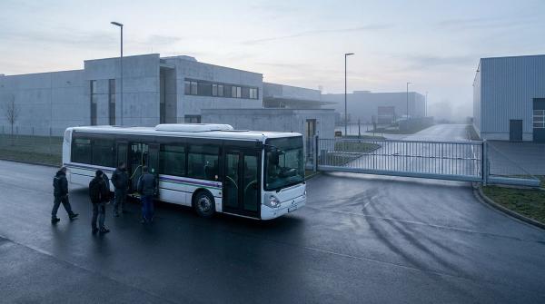 Beschäftigte steigen frühmorgens in einen Werksbus vor einem Werkstor im Gewerbegebiet, kein Linienbus in Sicht