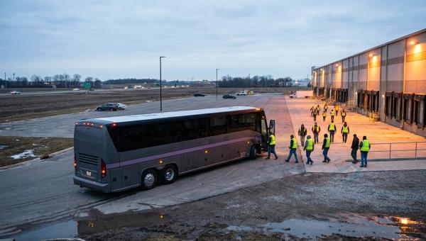 Workers in hi-vis vests walk from a parked coach toward lit loading docks at an exurban distribution center before dawn