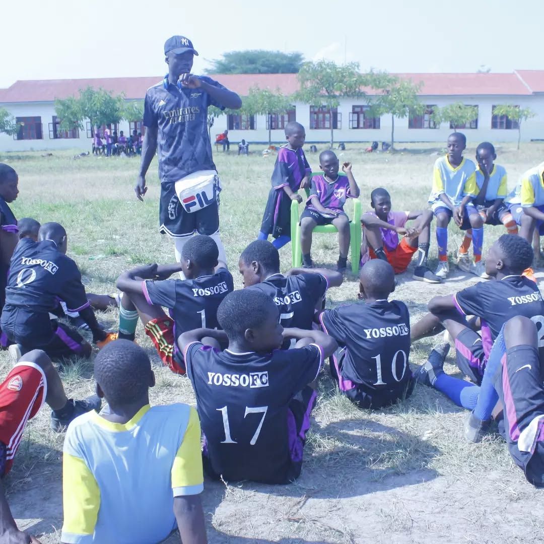 Coach speaking to Yosso FC players seated on grass in front of a school building