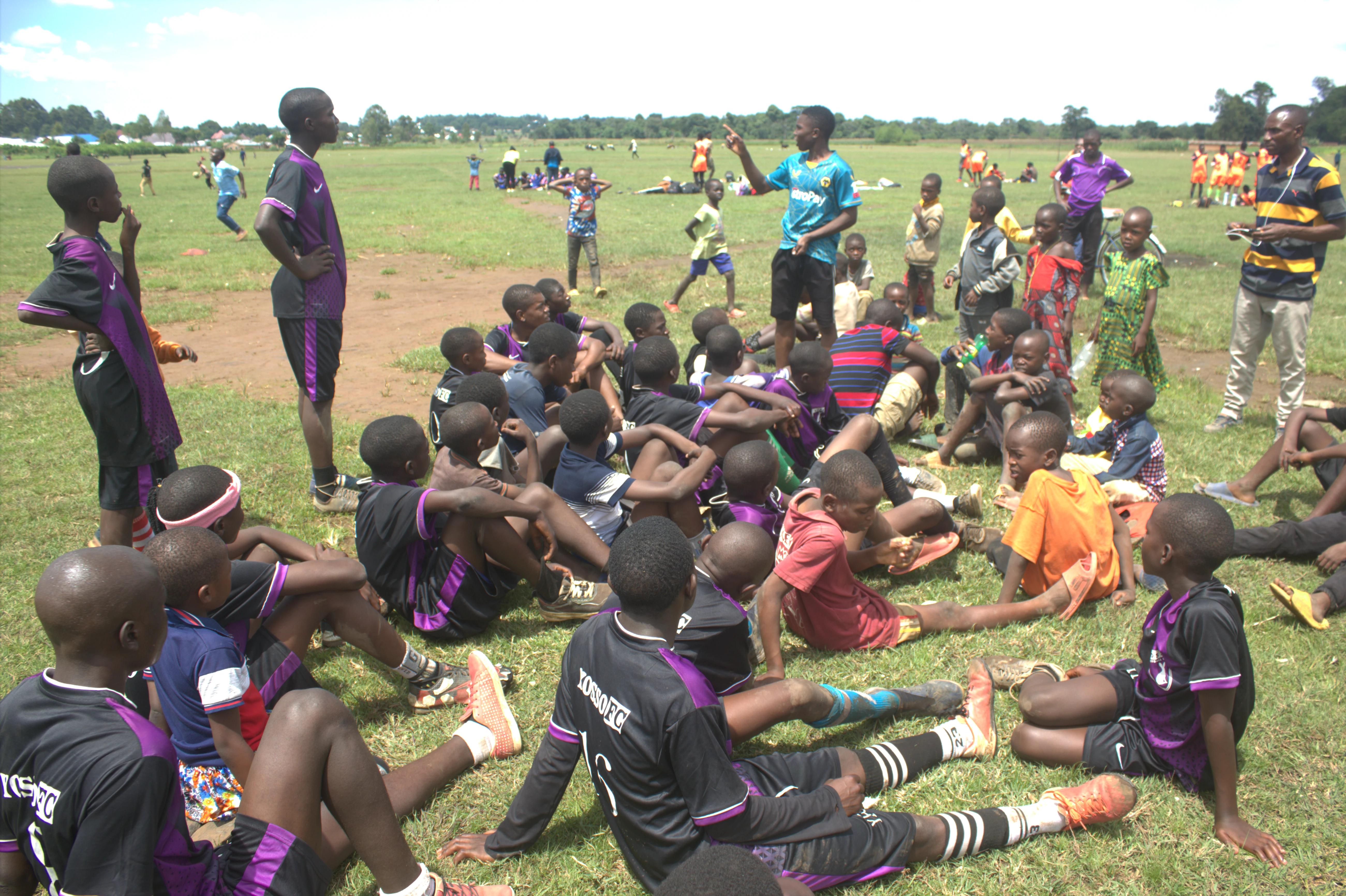Coach speaking to Yosso FC players seated on the grass during training