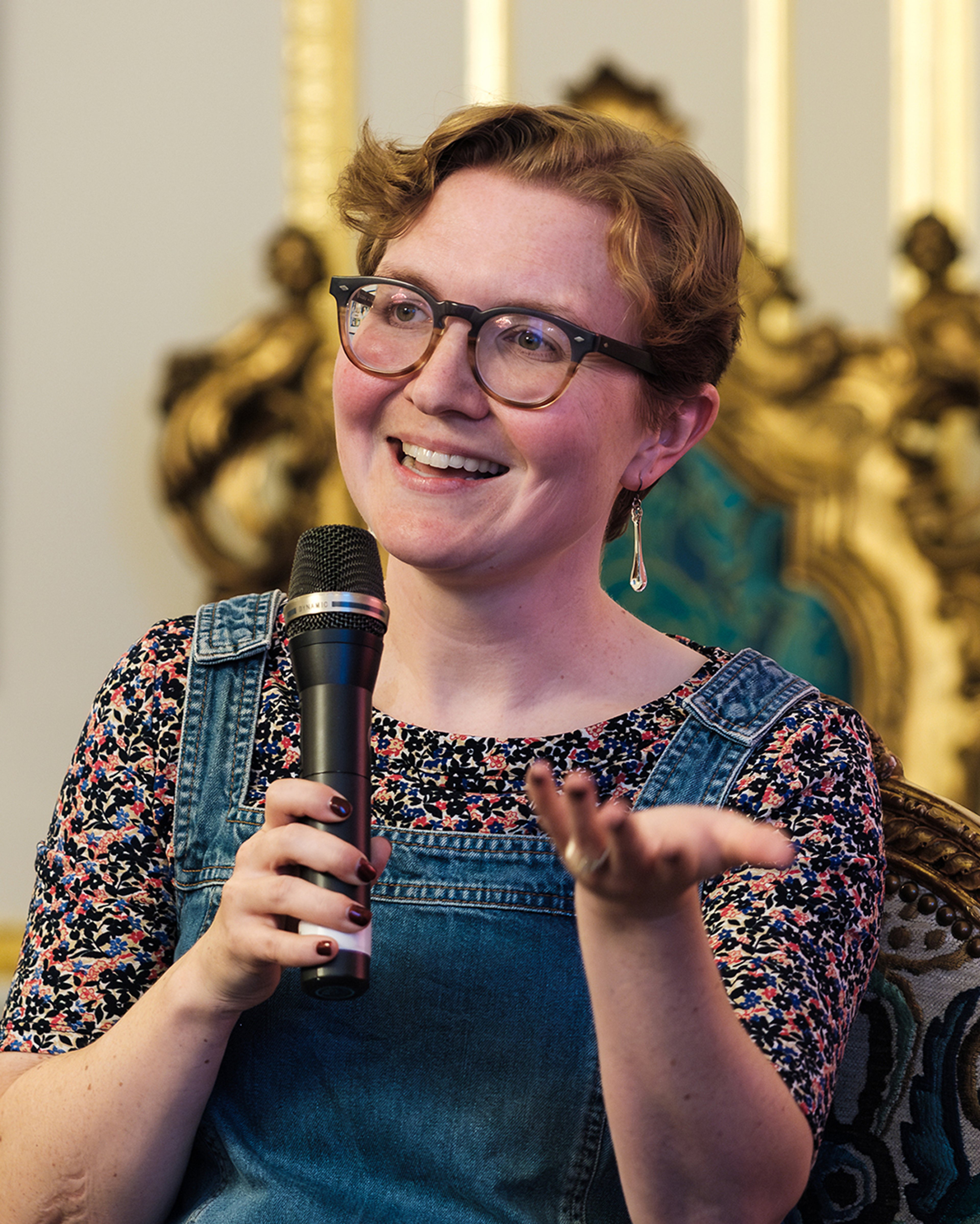 Katie, a white woman with short blonde hair, is smiling and talking to a crown (out of frame) using a microphone. She is wearing a flowery top and denim dungarees. She is sat on an ornate gold chair at the Canadian Embassy!