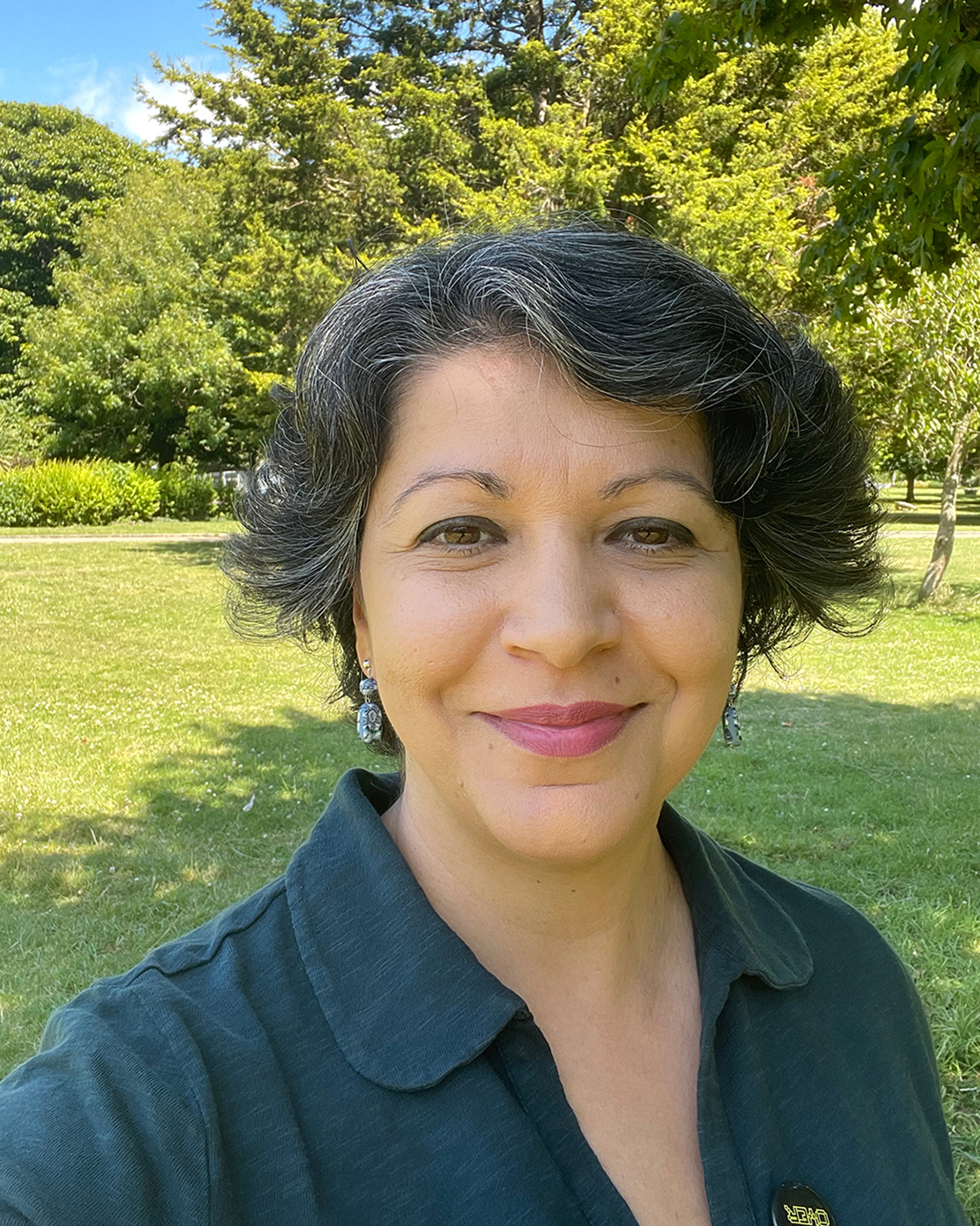 Portrait of Sana, an Iraqi woman, outside amongst the trees. She is smiling and wears a green top and pink lipstick.