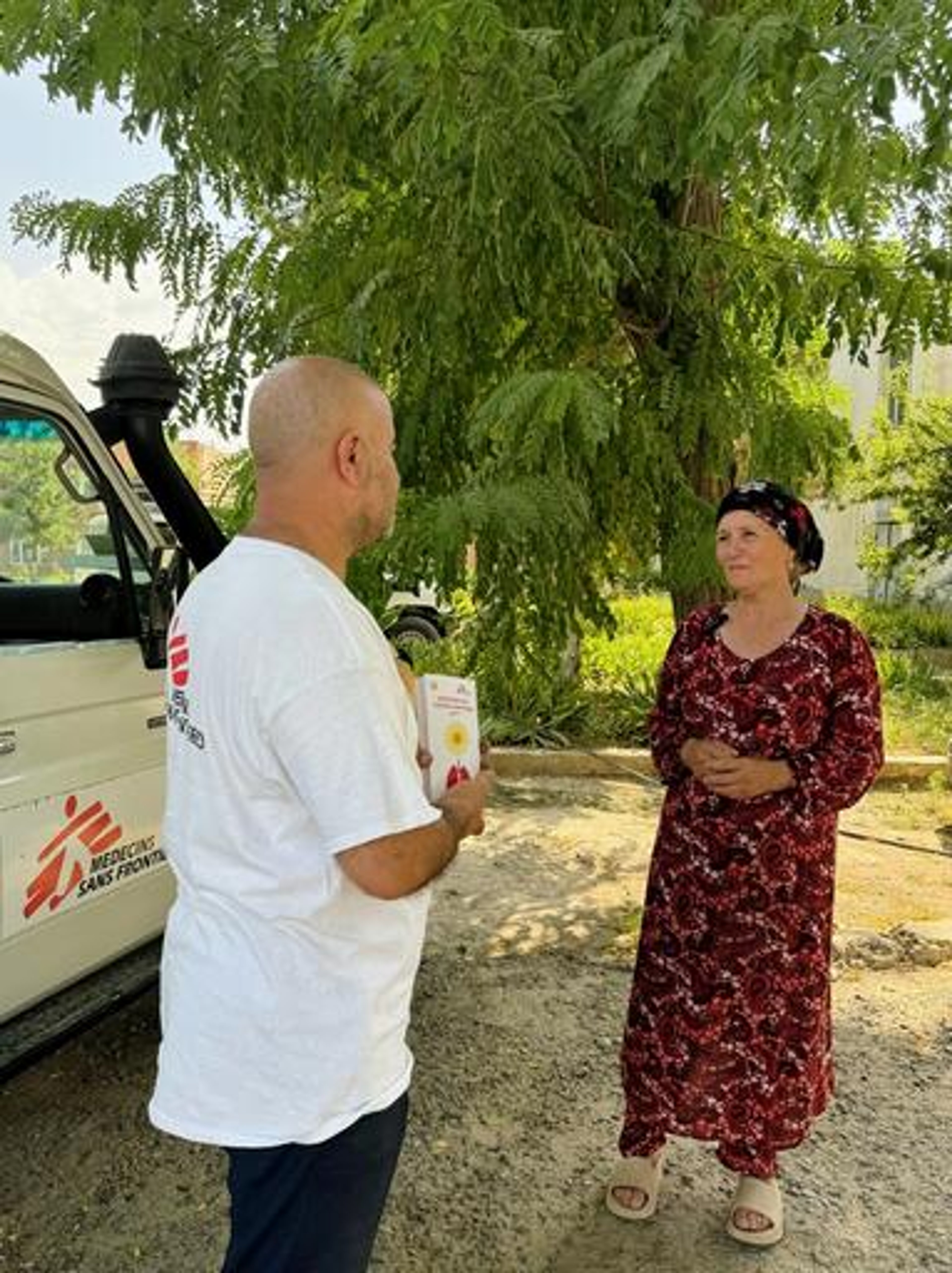 Sokinamo Khudoiberdieva, 59, from Kulob, southwestern Tajikistan, speaks with Askar Usmonov, MSF’s health promotion officer, before her X-ray. They are outside under a tree and stood by a white car with the Medecin Sans Frontieres logo on the door.
