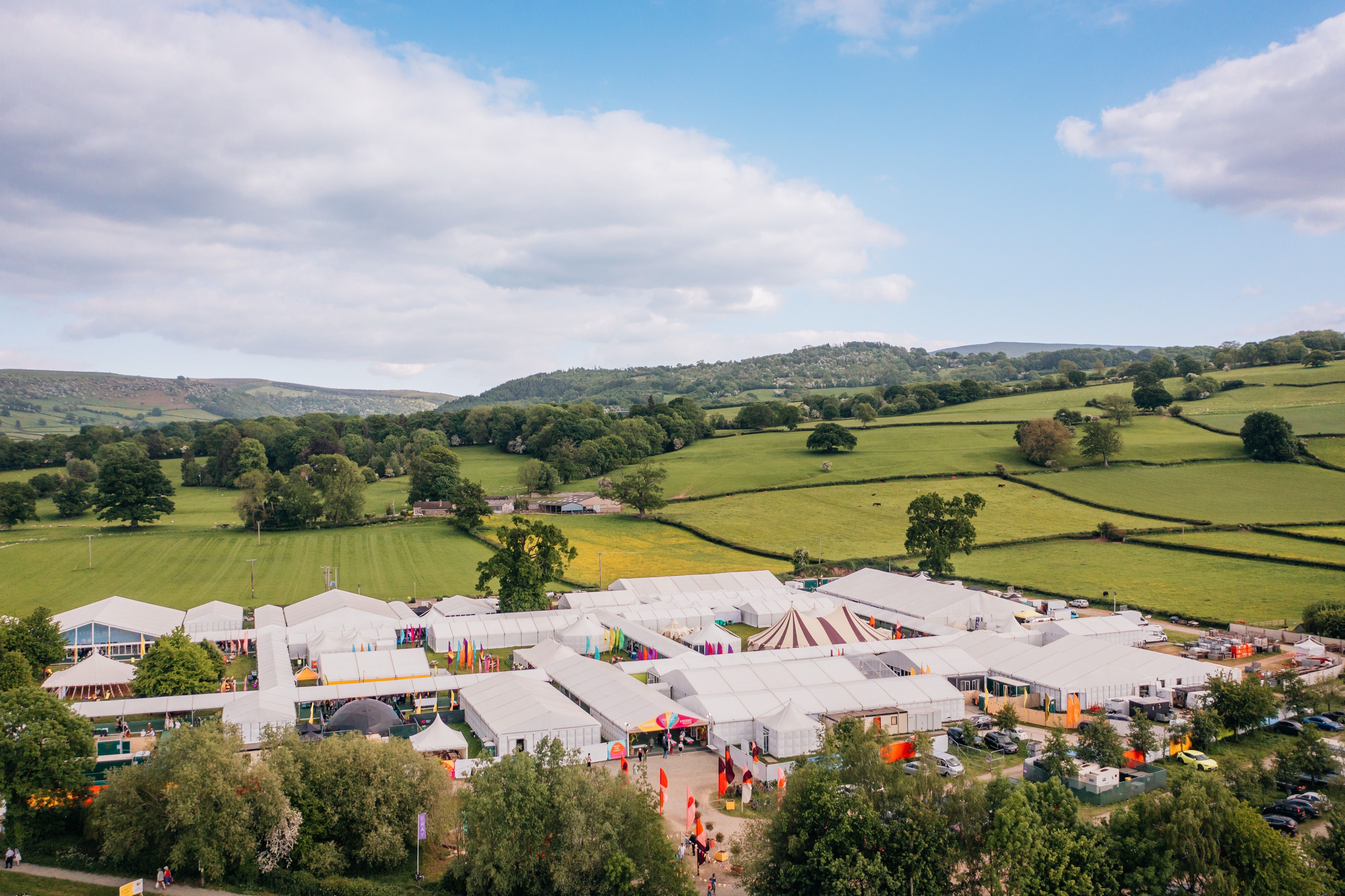 Image of Hay Festival in the Welsh countryside. A complex of white tents and colourful flags are in the foreground, and the rolling hills of Hay-on-Wye in the background.