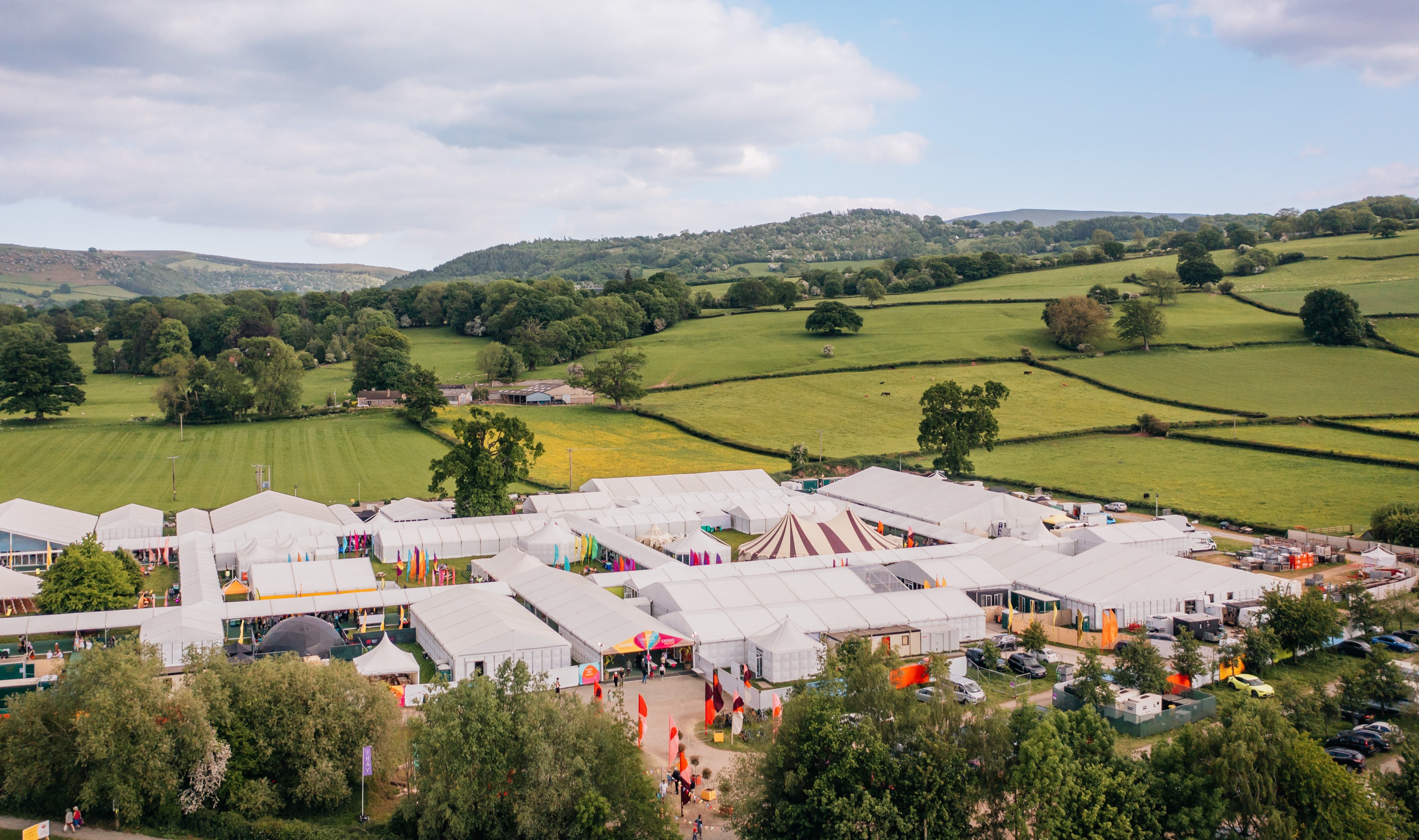 Image of Hay Festival in the Welsh countryside. A complex of white tents and colourful flags are in the foreground, and the rolling hills of Hay-on-Wye in the background.