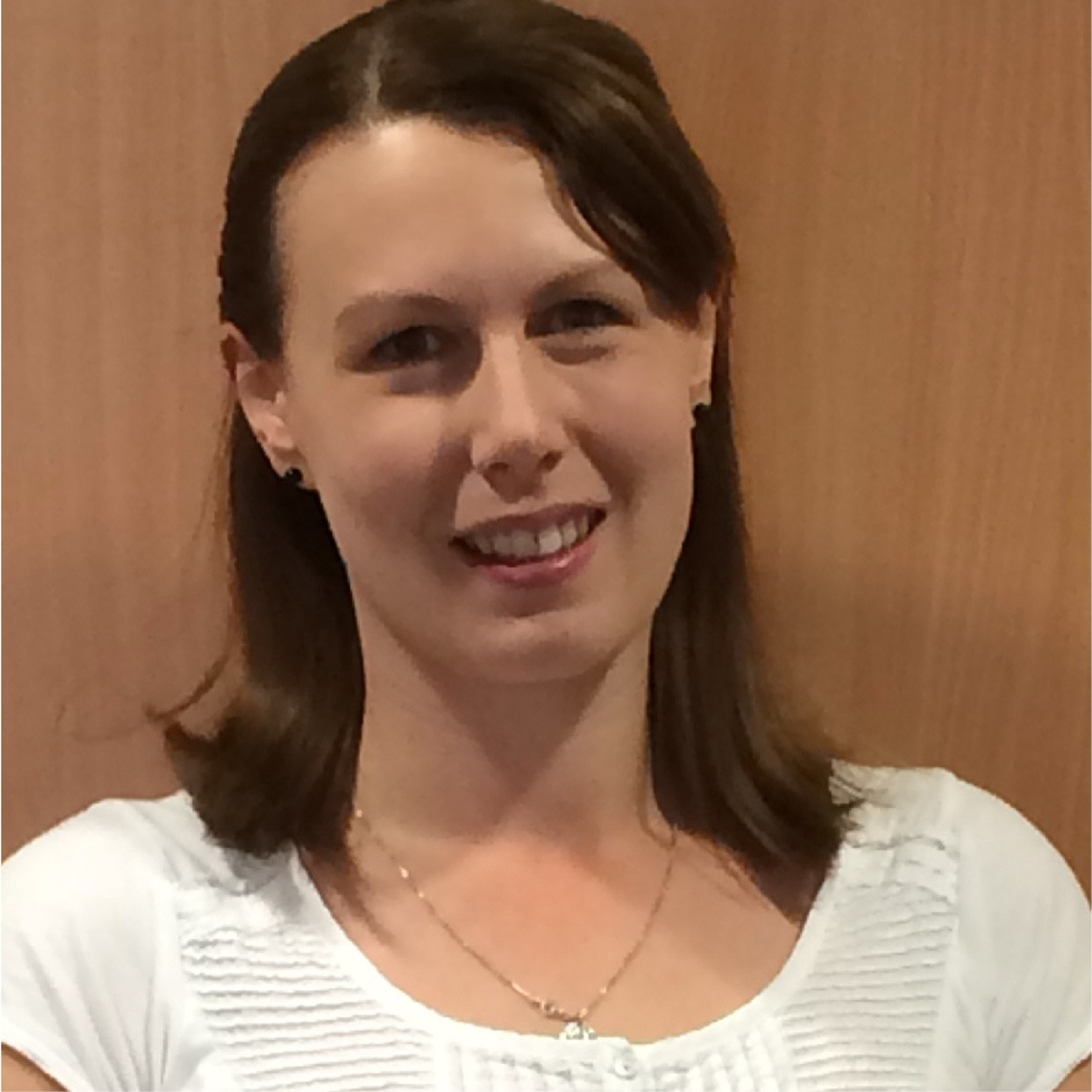 Portrait of Caroline, a smiling white woman with mid-length brown hair. She wears a white top and stands against a plain wooden background.