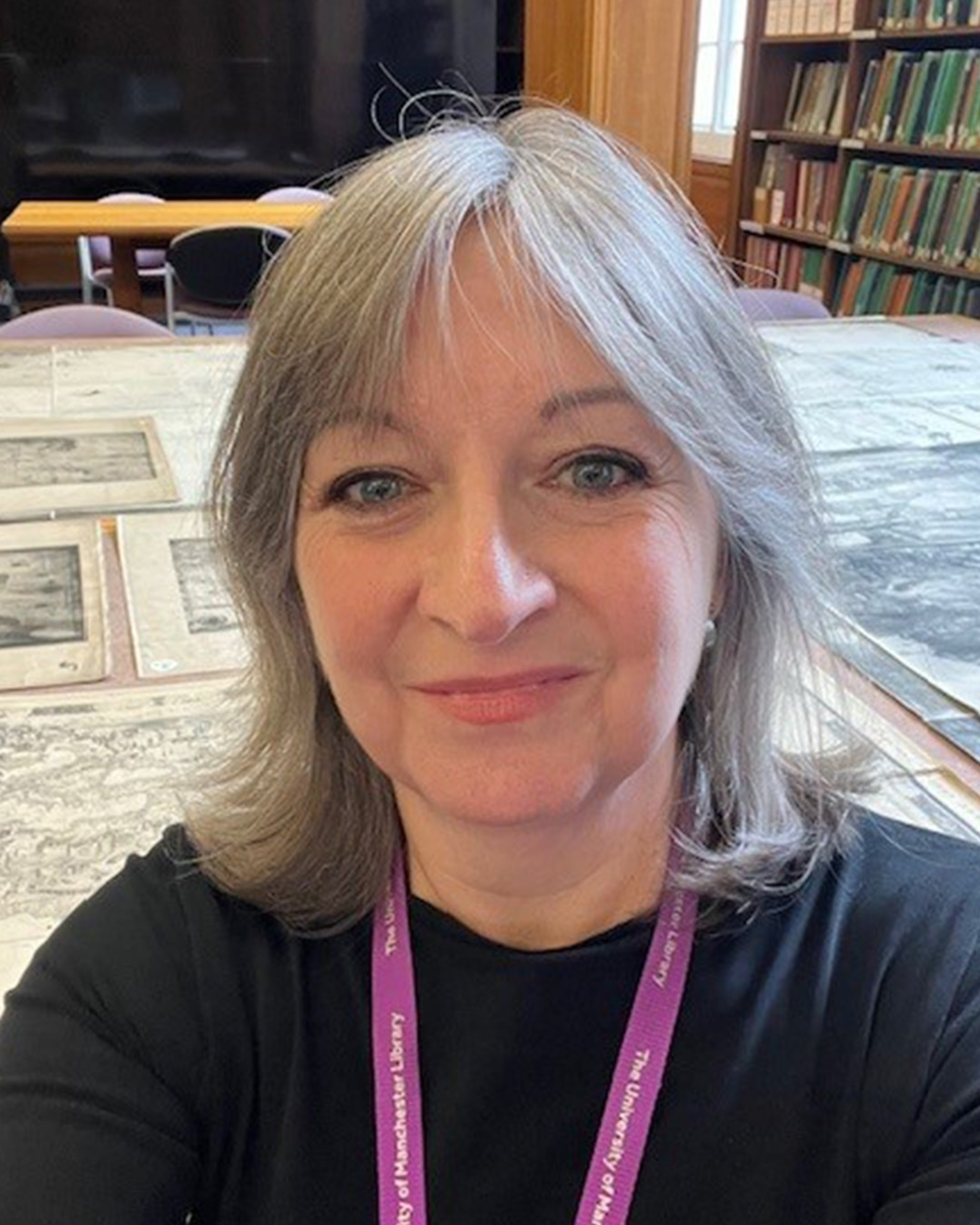 Headshot portrait of Donna, a white woman with fair mid-length hair and blue eyes. She is wearing a black top and a purple lanyard around her neck.  She is smiling and taking her self-portrait in front of a library table full of old maps, and with bookshelves in the background.