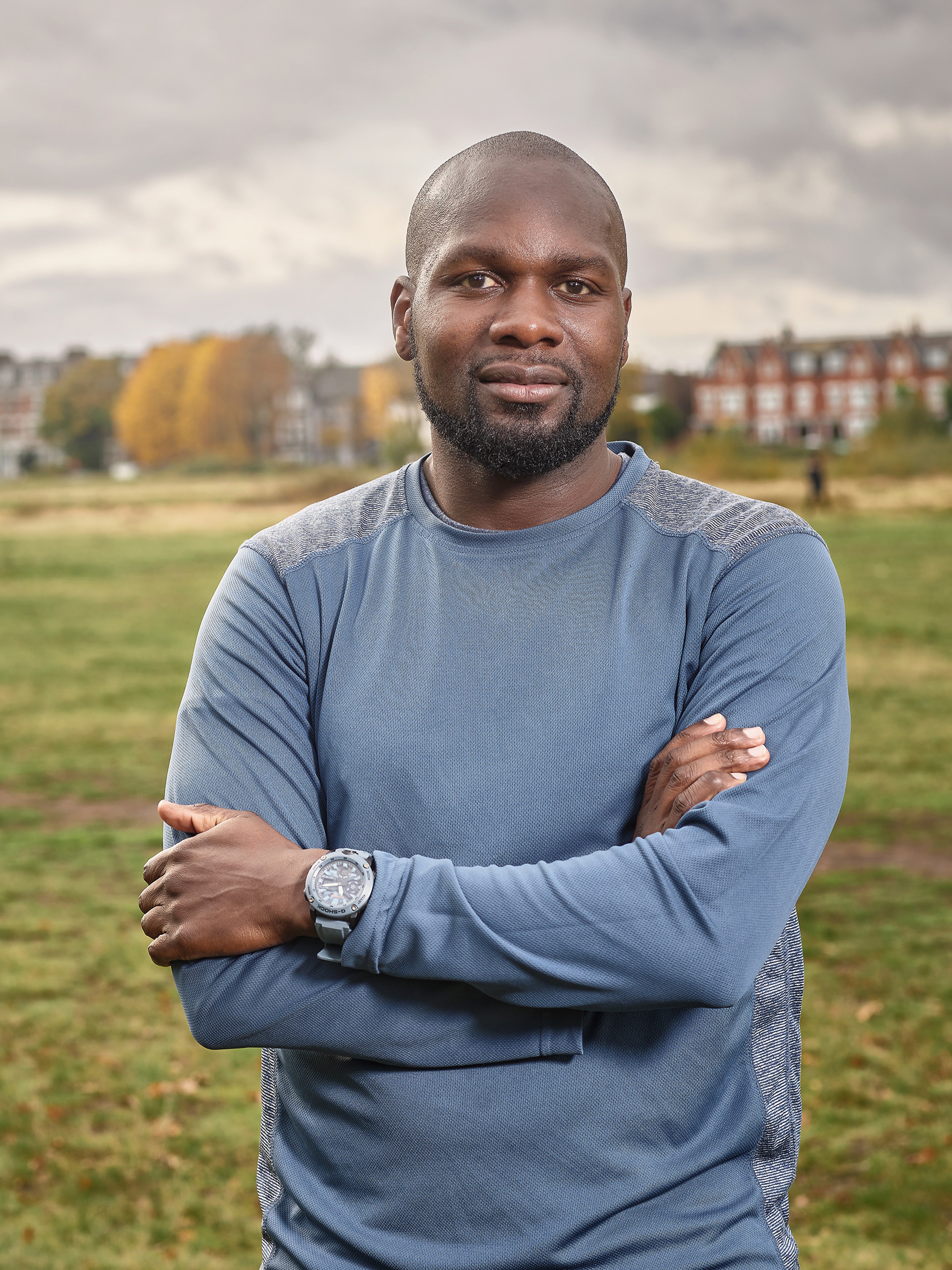 Portrait of Dwayne, an athletic black gentleman with a beard and a shaved head wearing a long-sleeve blue top. He stands outside in green space with houses in the background