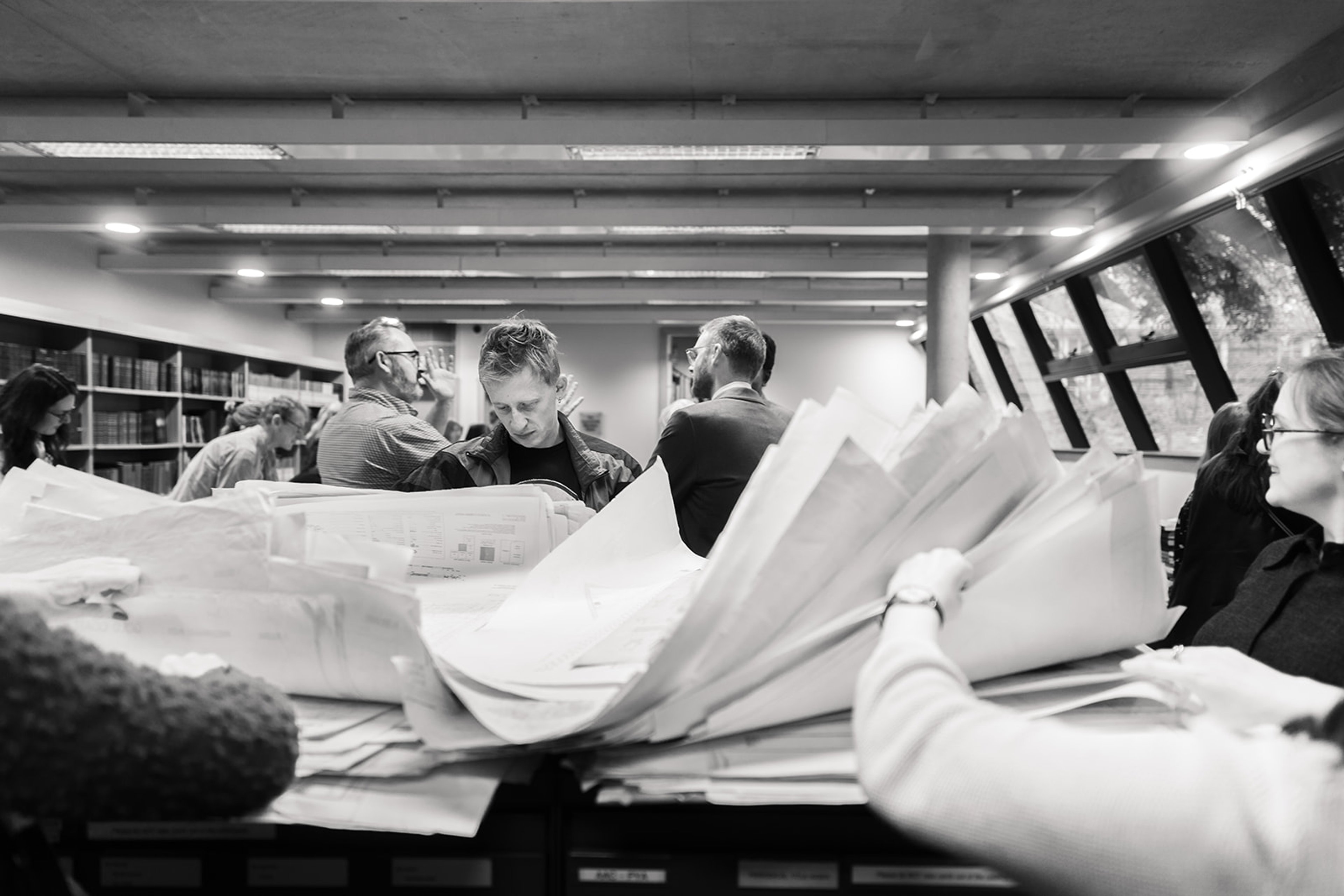Black and white photograph of busy RGS reading room and people looking through piles of maps.