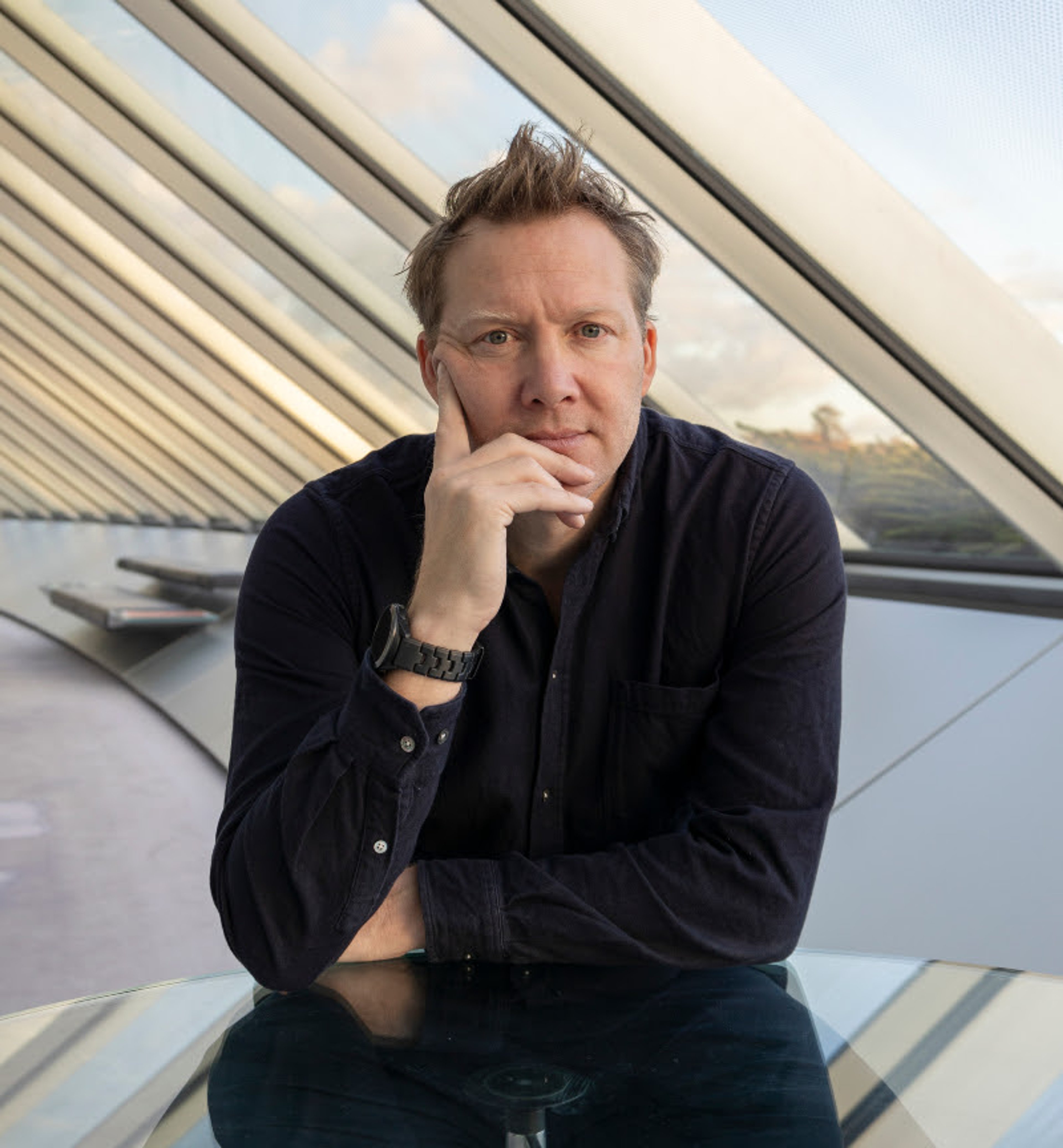 Portrait of Jago Cooper, a white man who is sat a table and resting his face on his hand. His curious expression as if asking a question, or listening to an answer. He has short spikey hair and a black shirt on. He is sat in a space with lots of windows and the light is very atmospheric.