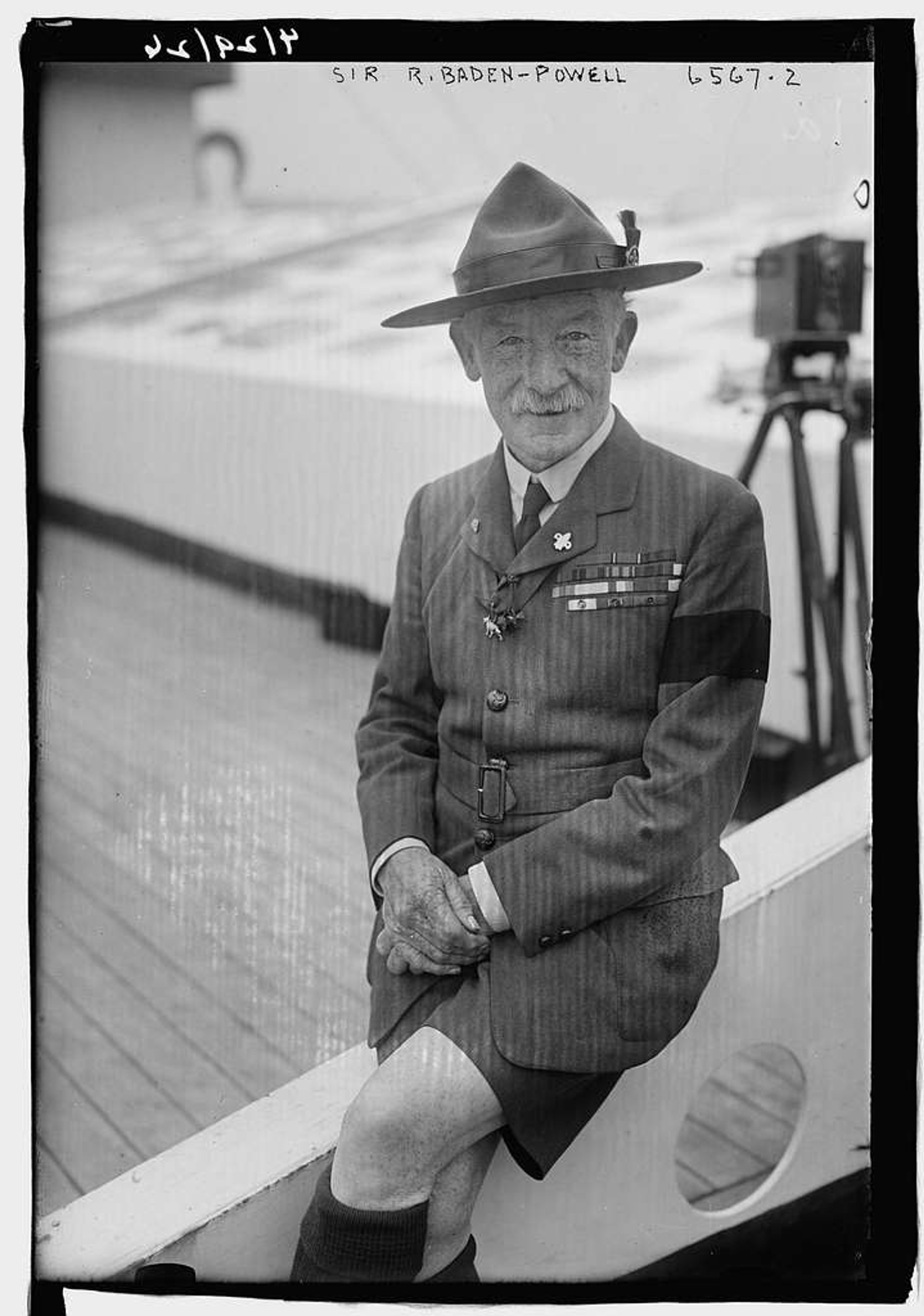 Black and white portrait of Scout movement leader Baden-Powell. He is sat on a wall or railing smiling directly at the camera. He is wearing a hat, and his military uniform and shorts with high socks.
