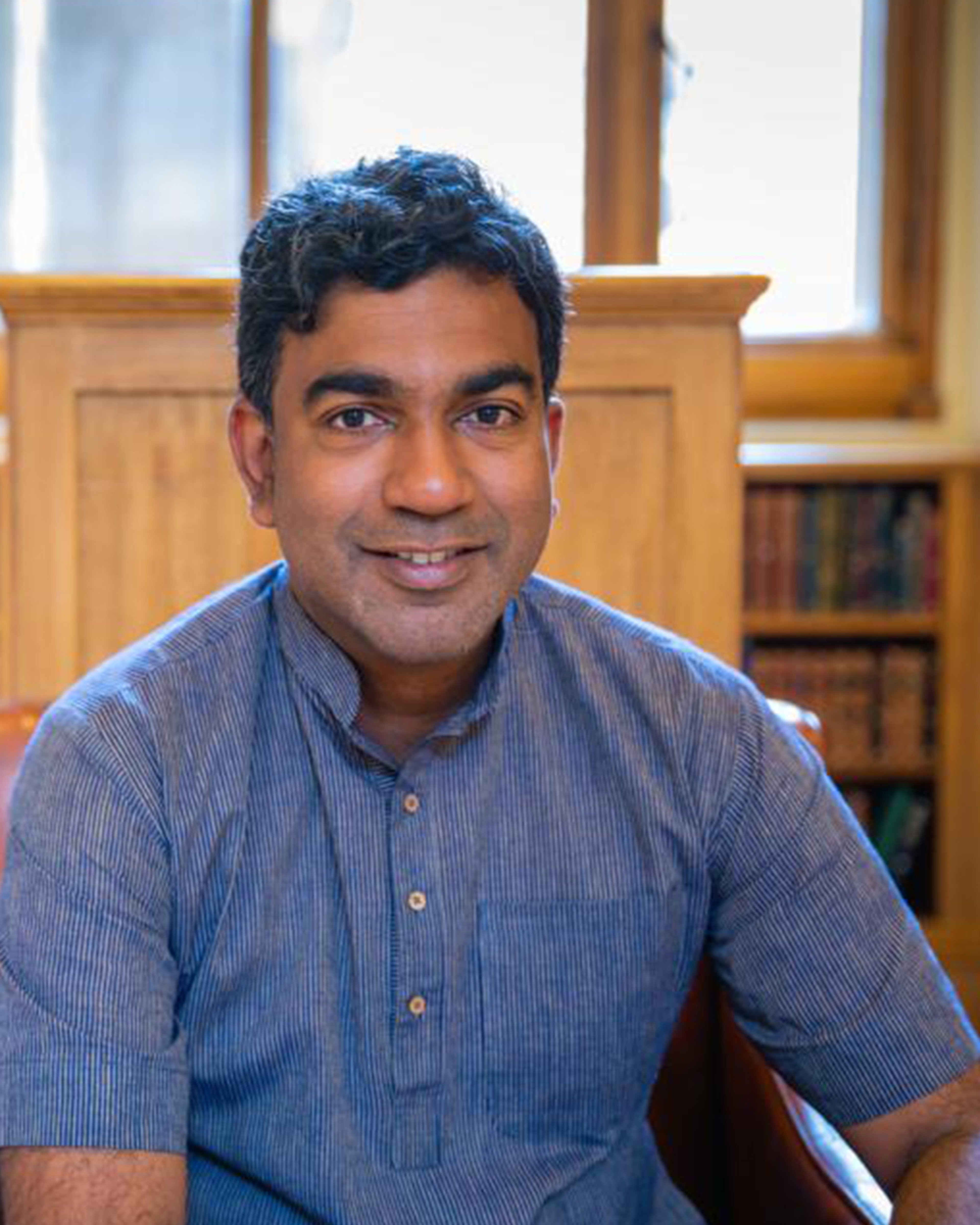 Portrait of a Sri Lankan man wearing a blue shirt, sat in a wood-panelled library environment, with booked in the background. He is smiling.