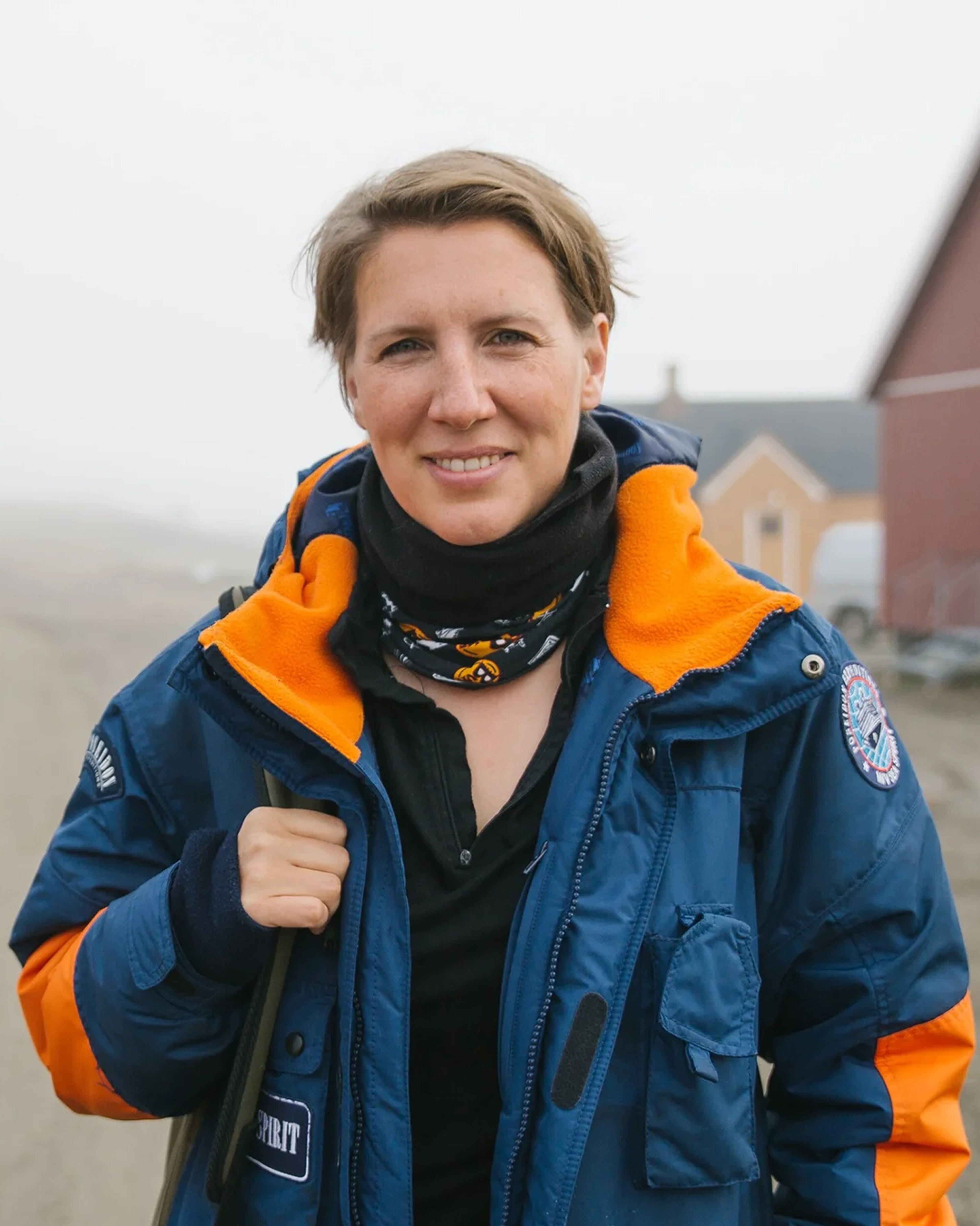Portrait of Felicity, a white woman with short brown hair. She is smiling and wearing a big blue and orange expedition jacket/coat, and holding her backpack on her shoulder. She is outside in a misty location with some houses in the background.