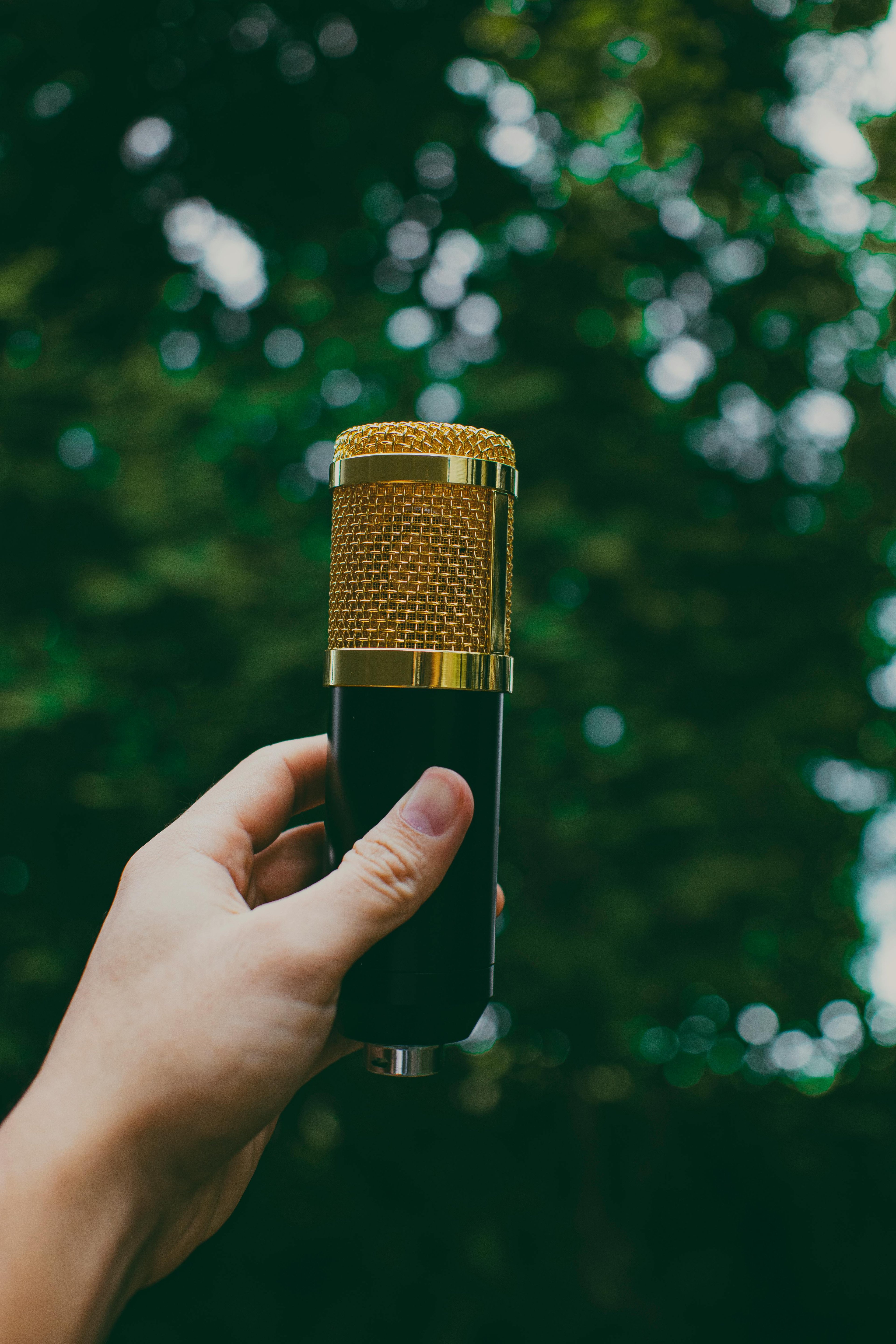 Soft focus image of a white hand holding a gold microphone up against green foliage of tree / to record nature