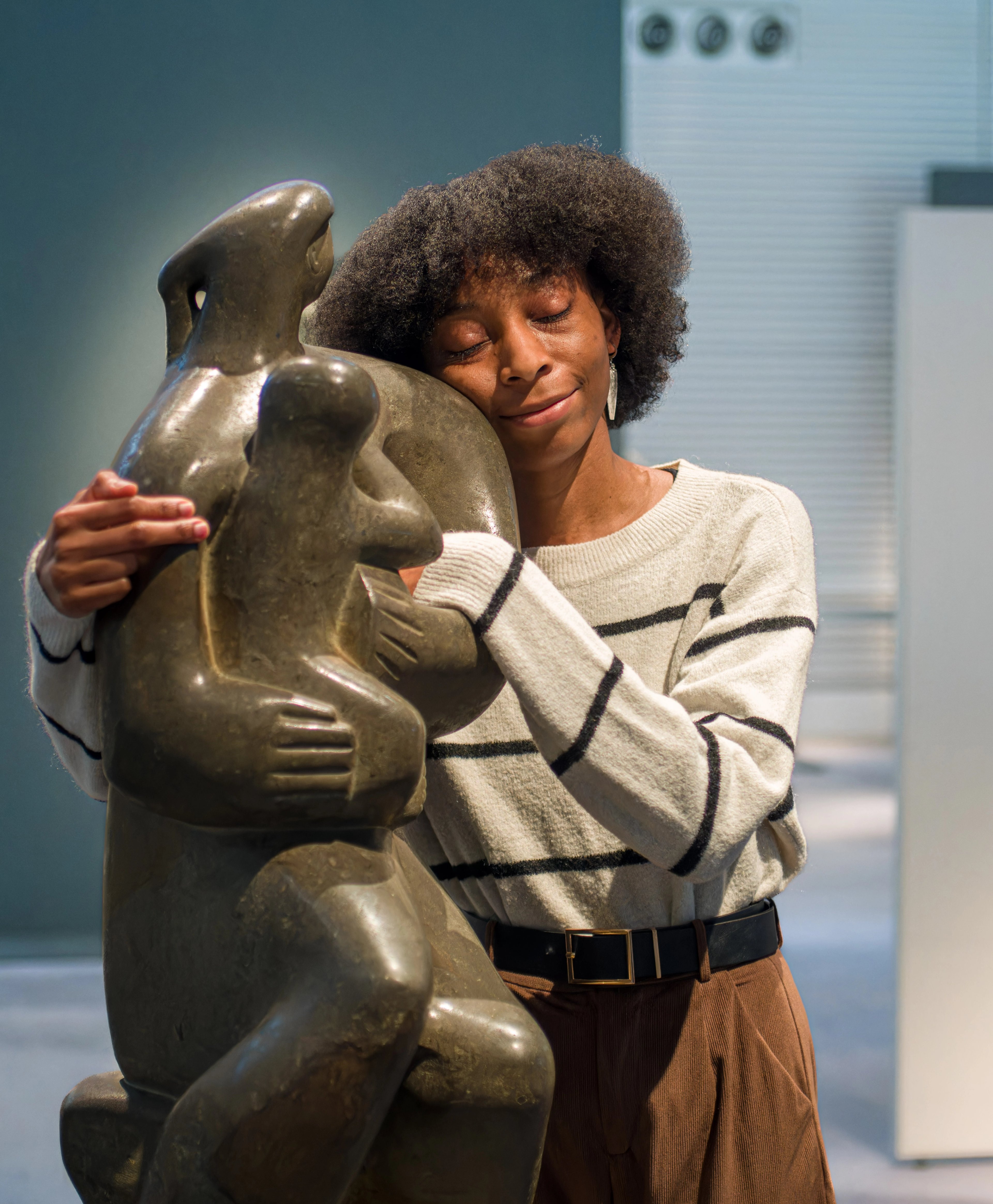 A black woman wearing a white striped jumper and brown trousers is hugging a Henry Moore sculpture 'Mother and Child' in a museum. She is smiling and has her eyes closed in a contented embrace.