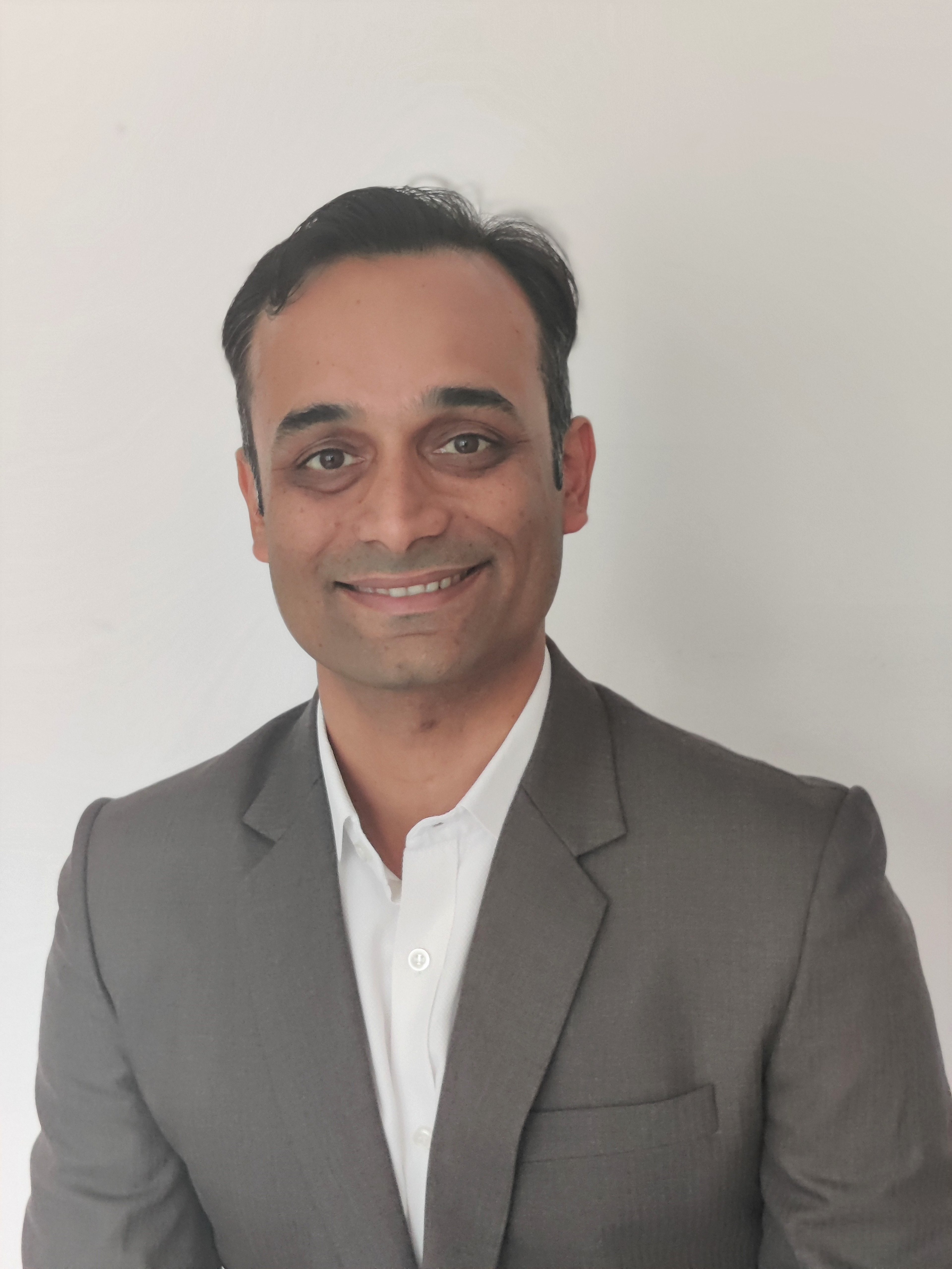 Portrait of Animesh Sinha, an Indian man who is smiling with kind eyes into the camera. He is wearing a smart grey suit and bright white shirt.