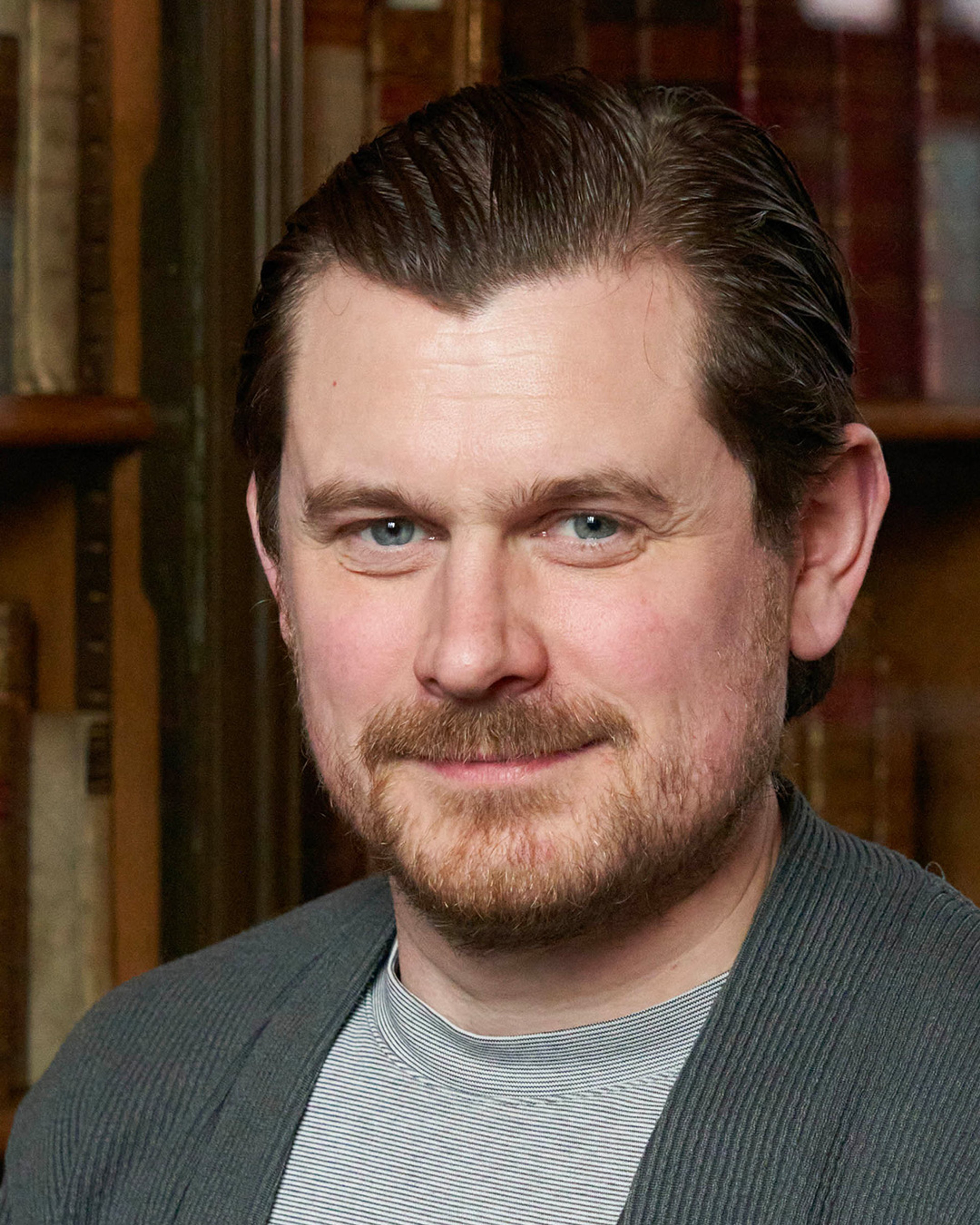 Portrait of Jamie Robinson, a white man with short brown hair and a beard. He is wearing a light shirt and a grey cardigan. He is standing in front of a bookshelf filled with old books.