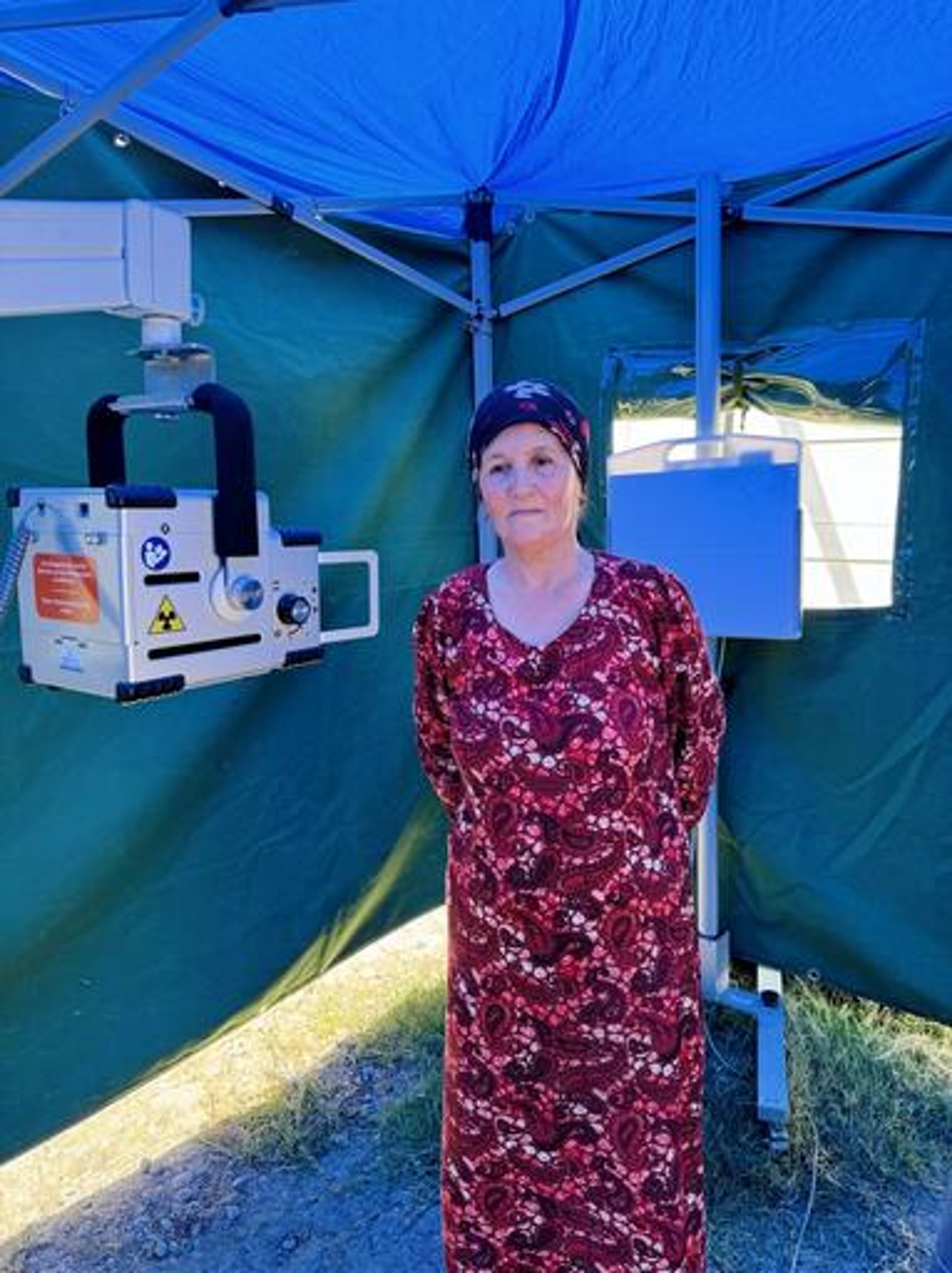 Sokinamo Khudoiberdieva, 59, from Kulob, Tajikistan, is preparing for her X-ray.. She is an older white woman wearing a long floral red dress and a headscarf. She is stood in a tent next to an x-ray machine.