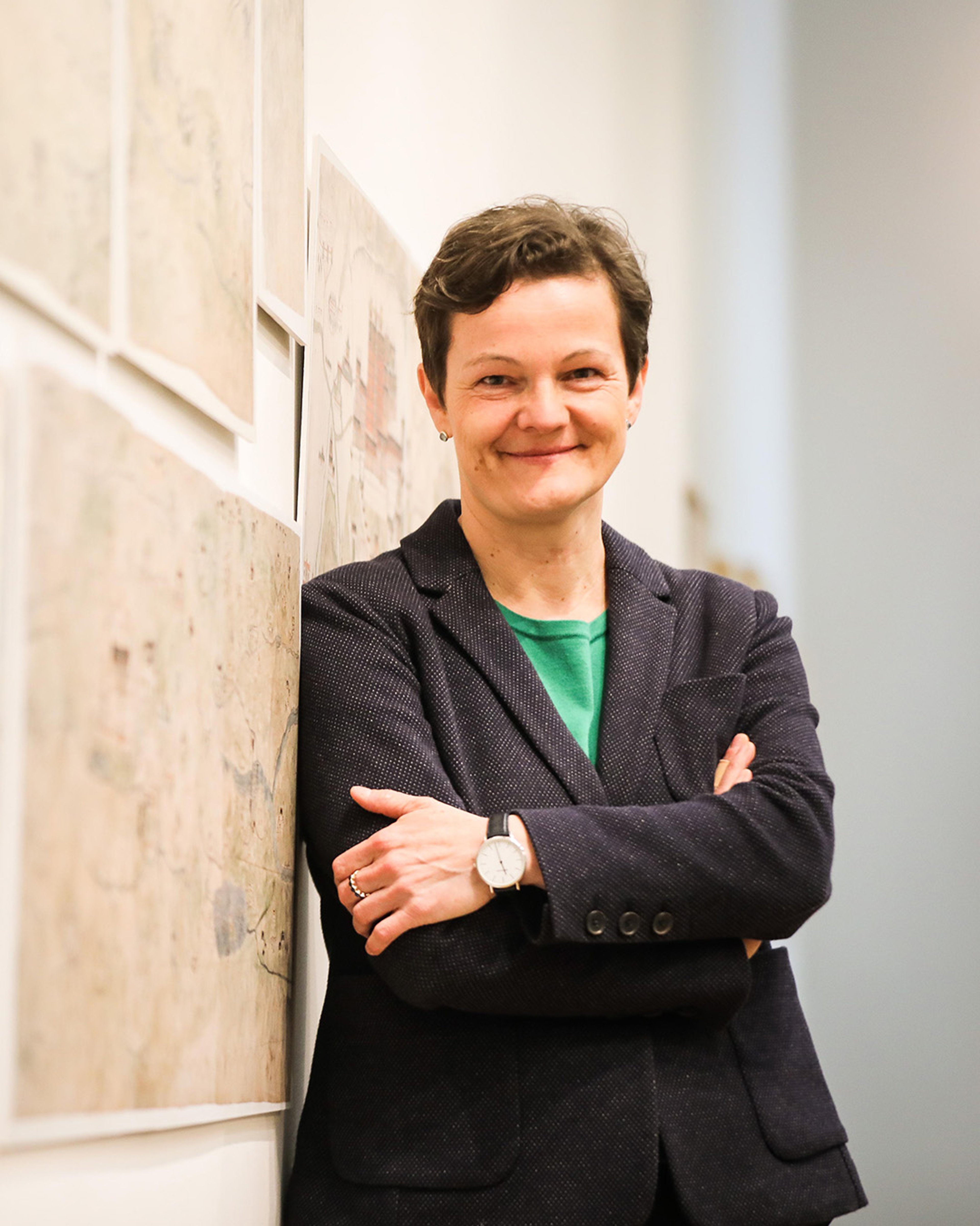 Portrait of Diana Lange, a smiling white woman with short brown hair. She is wearing a dark grey woven suit, a bright green shirt and a smart watch. She is leant against a wall with a display of old maps.