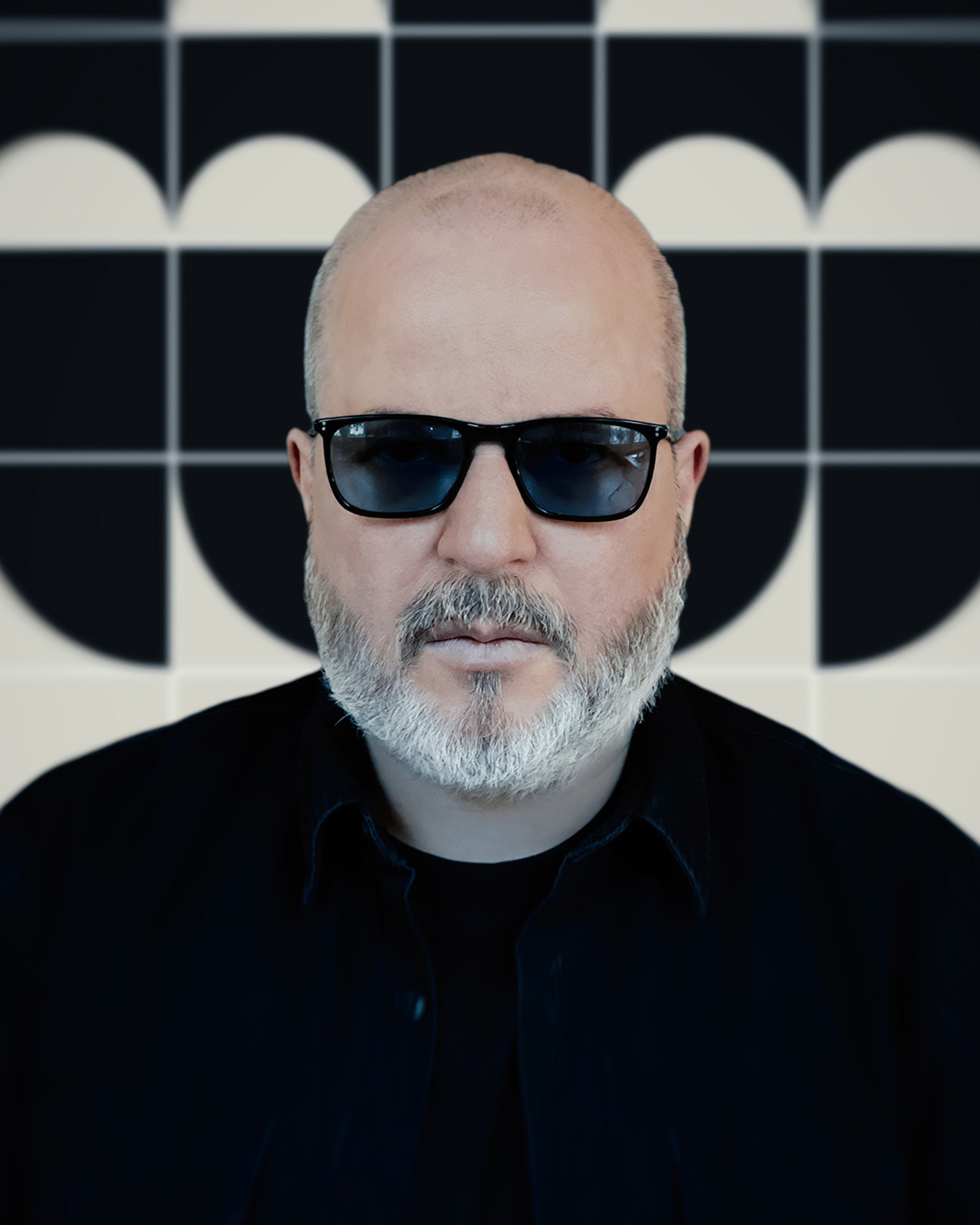 Portrait of Eric, a white man with a serious facial expression who is wearing cool dark sunglasses. He has a beard and is wearing a black shirt. He stands in front of a black and white patterned wall.