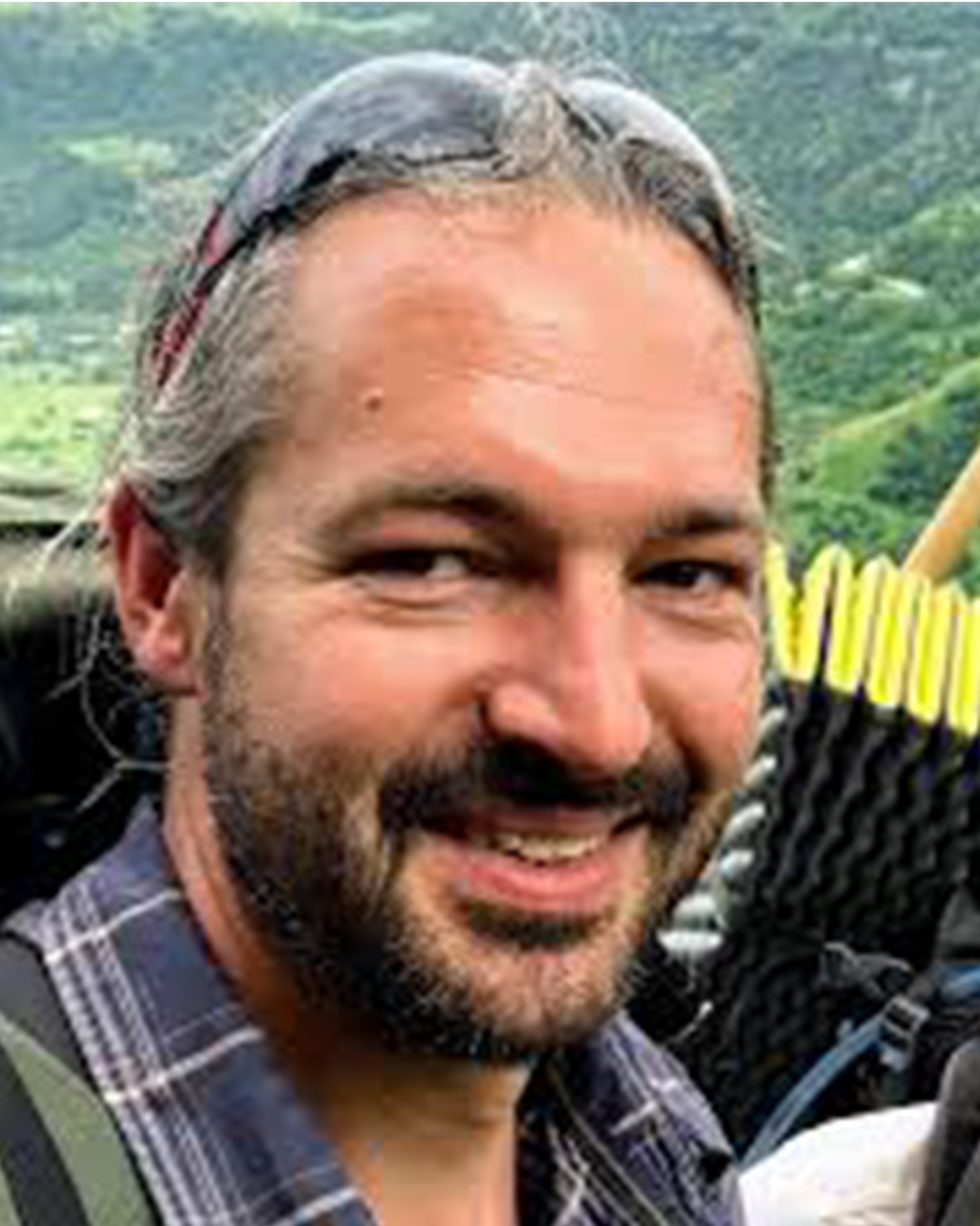 Tom, a white man with a beard and mousy brown hair, smiles at the camera. He is wearing a blue check shirt and sunglasses on his head. Just out of shot you can see the backpack he is wearing is overloaded with camping and hiking equipment.