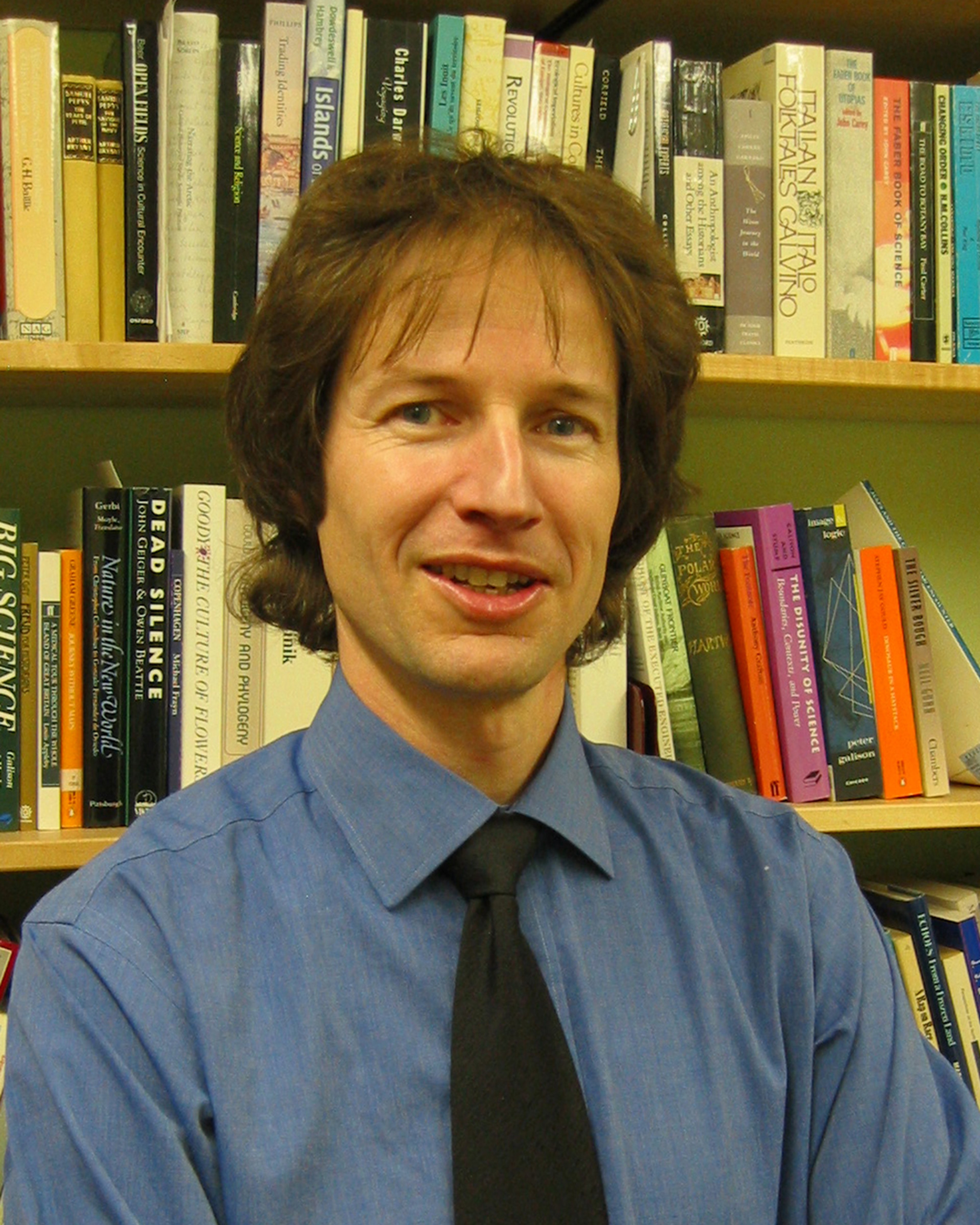 Michael, a white man with mid-length brown hair, is smiling and stoof infront of a bookshelf. He is wearing a blue shirt and a black tie.