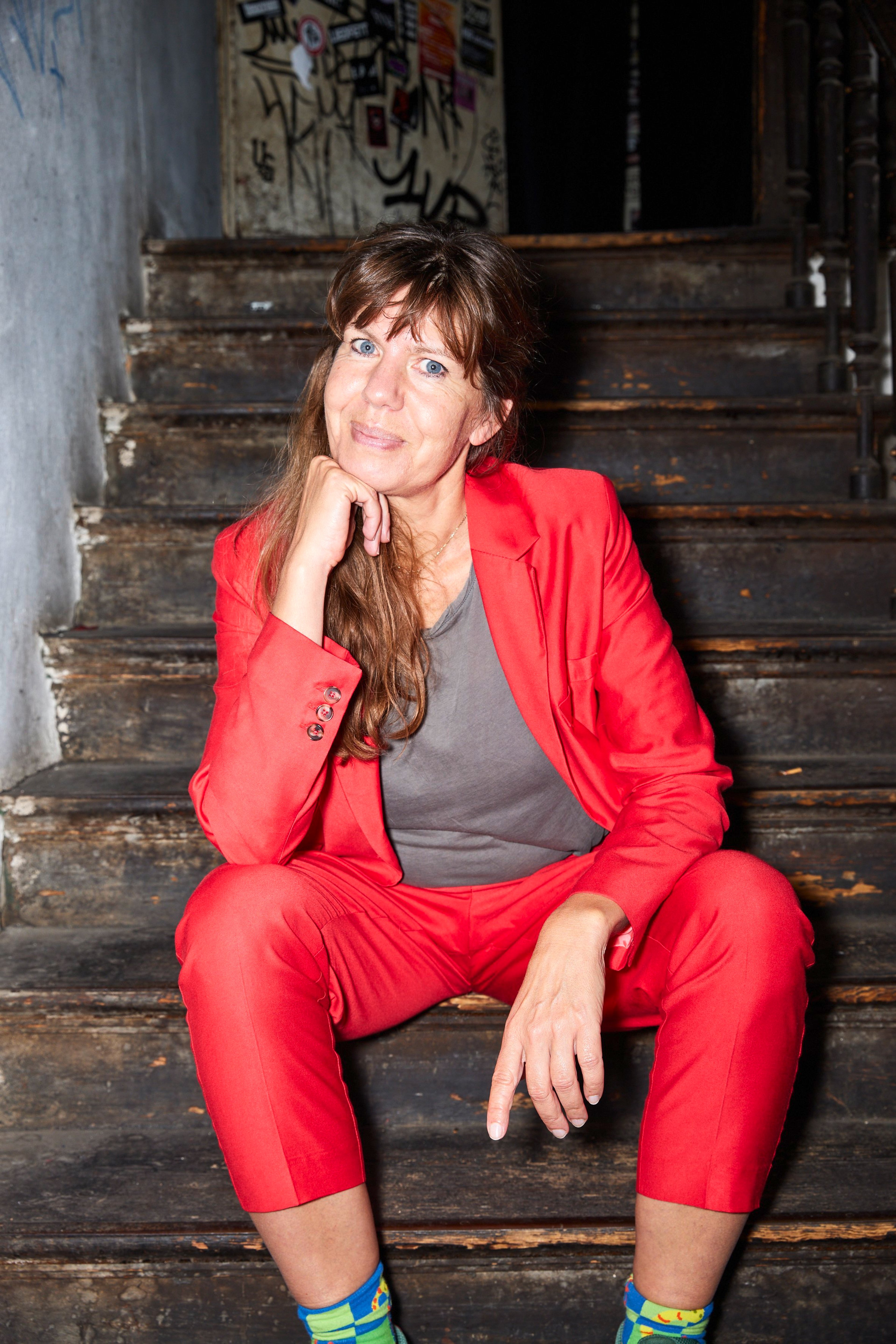Portrait of Michaela, a white woman wearing a bright red suit and a grey shirt. She is sat on wooden stairs and smiling at the camera.