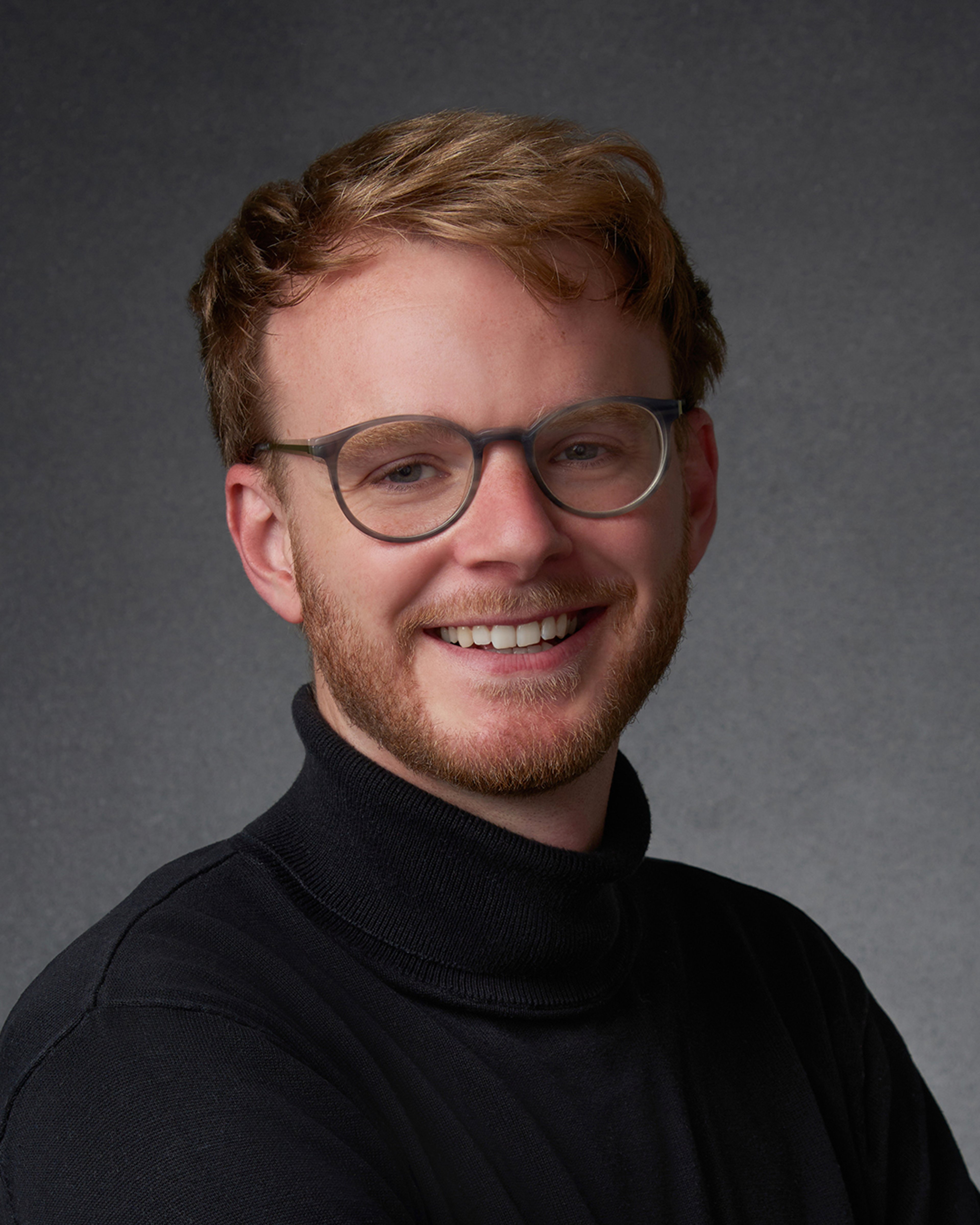 Portrait of a white man with ginger/fair hair, wearing a black turtle neck jumper. He is smiling and wearing glasses.