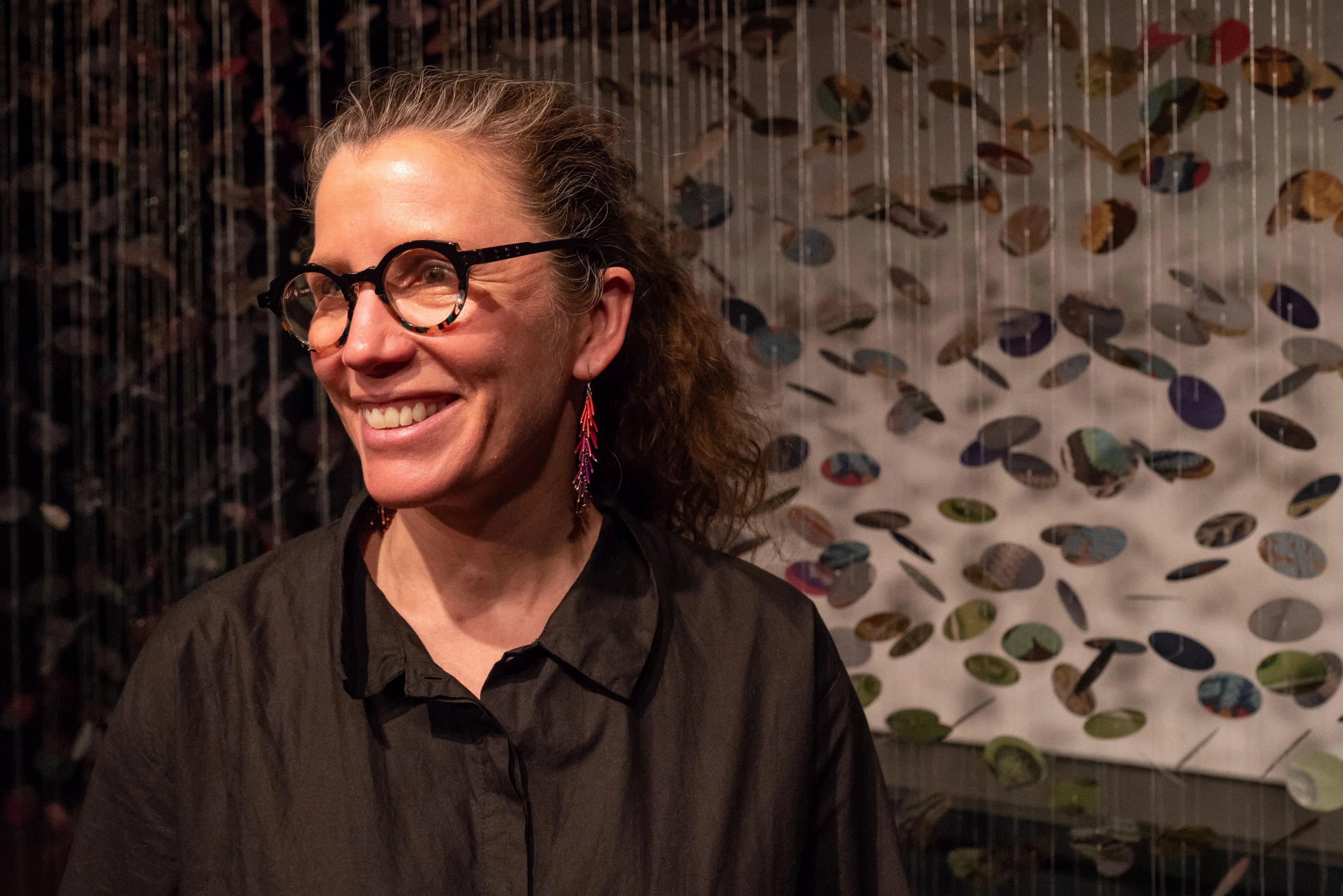 Portrait of Canadian First Nations artist Hannah Claus in front of her installation artwork. Hannah is smiling - she has mid-length wavy hair which is tied back. She wears a black shirt, glasses and colourful drop earrings. A light shines on her face.