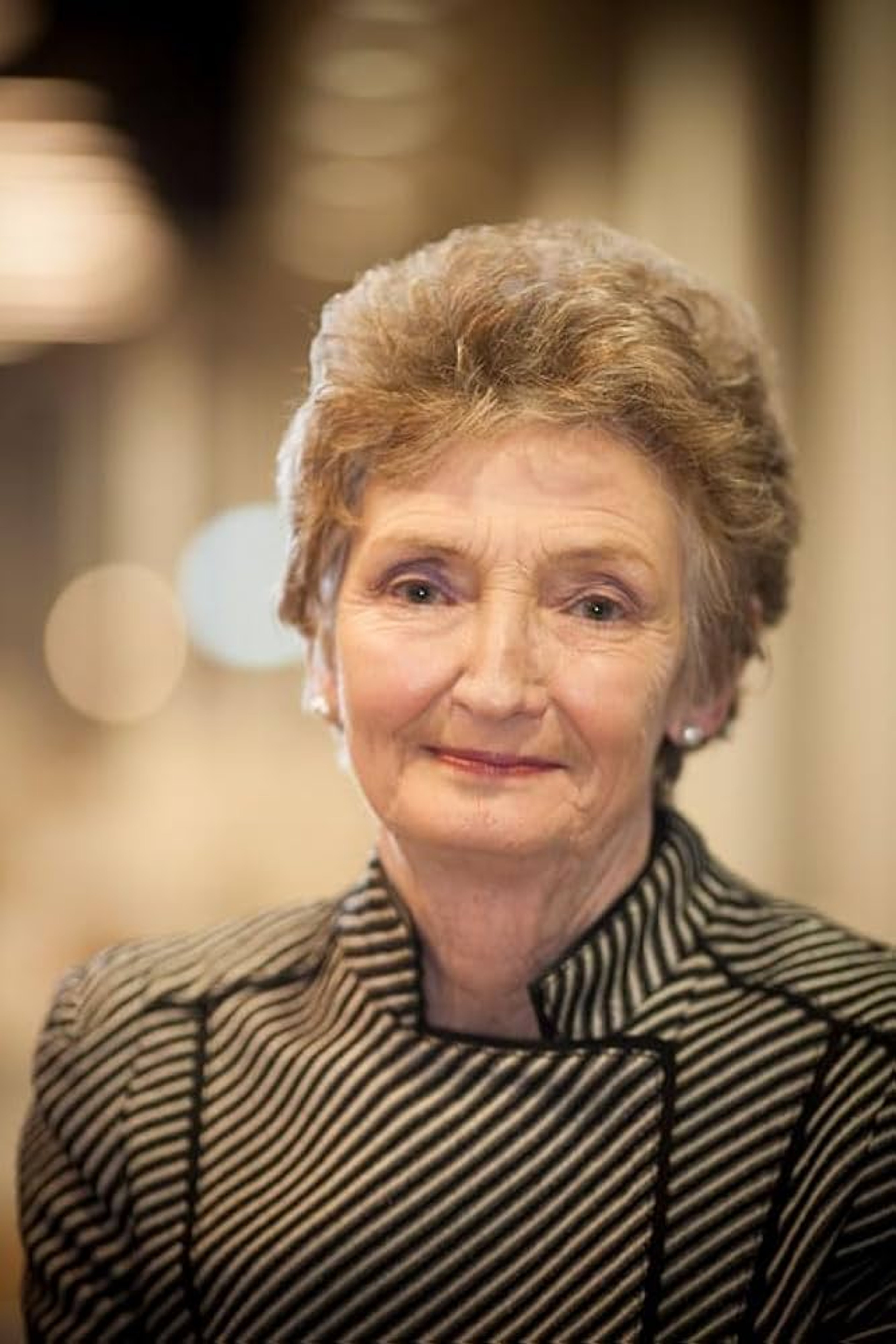 Portrait of Barbara, an older white lady who is has shorter blonde hair and red lipstick on. She is dressed smartly in a striped jacket and wears pearl earrings. She smiles at the camera, with a soft background of warm lighting.