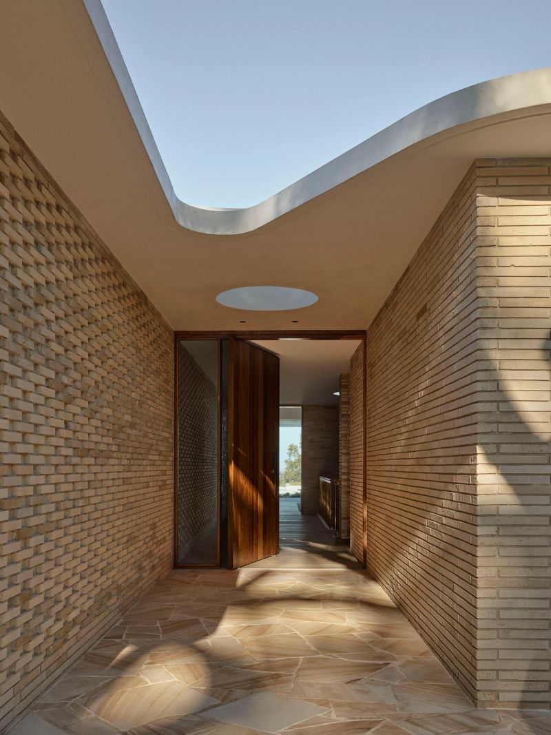 Entry passage at Breeze House between brick walls, under a curved white roof and round skylight, leading to an open timber pivot door