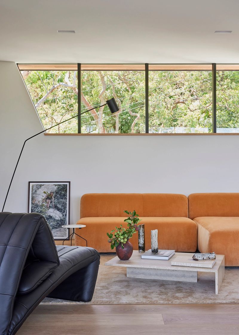 Living room at Cobbittee House with a tan sofa, black lounge chair and a high window to the trees