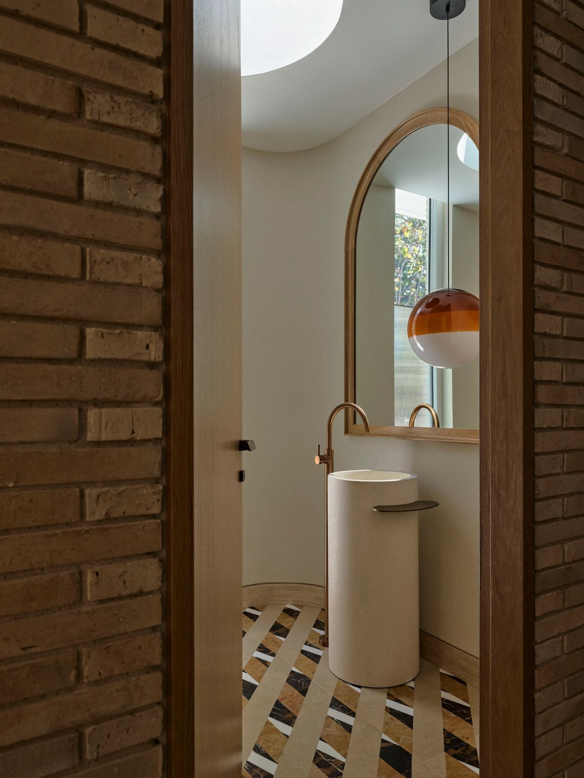 Powder room at Breeze House seen through a brick doorway, with a pedestal basin and arched mirror under a skylight