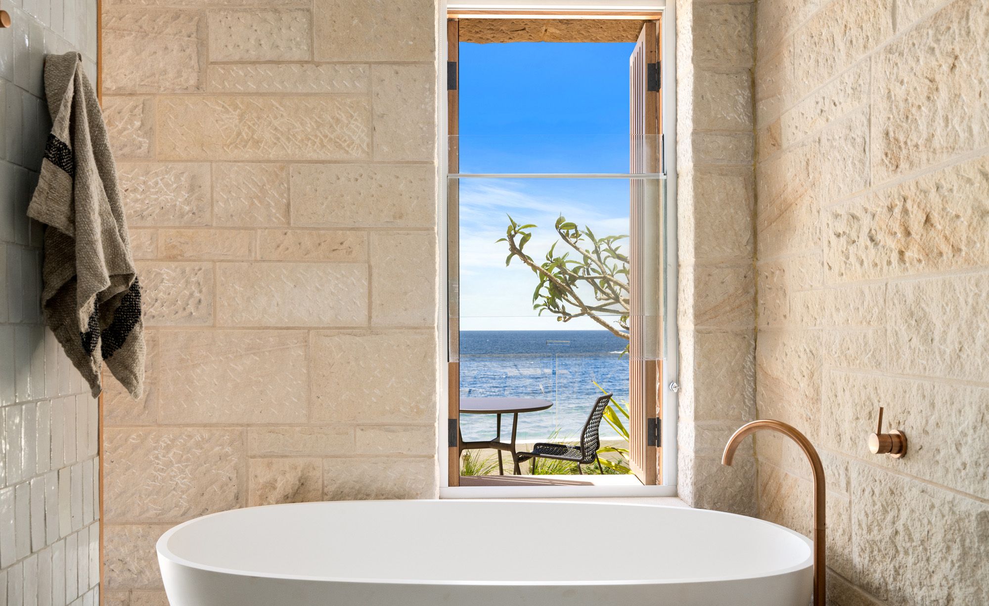 Freestanding bath in a sandstone bathroom with a window to the sea