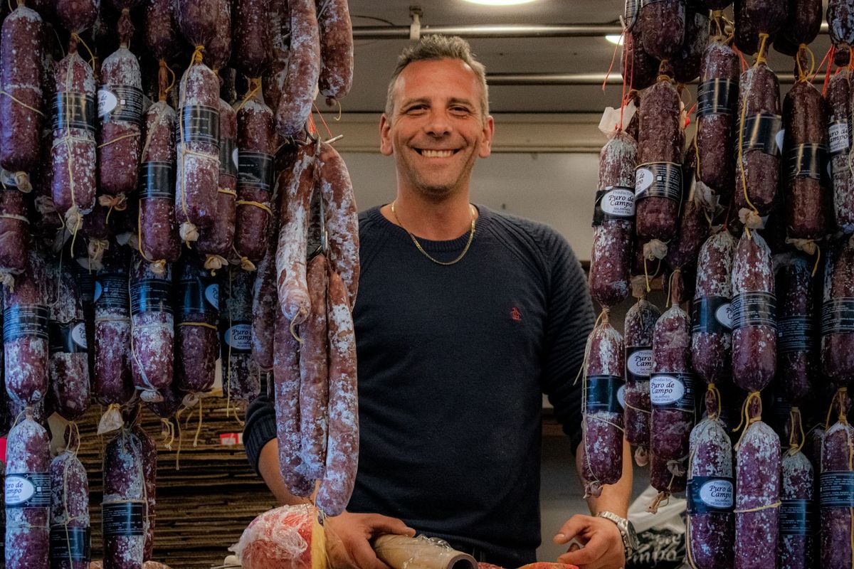 Vendeur souriant devant des salamis suspendus dans son étal de marché