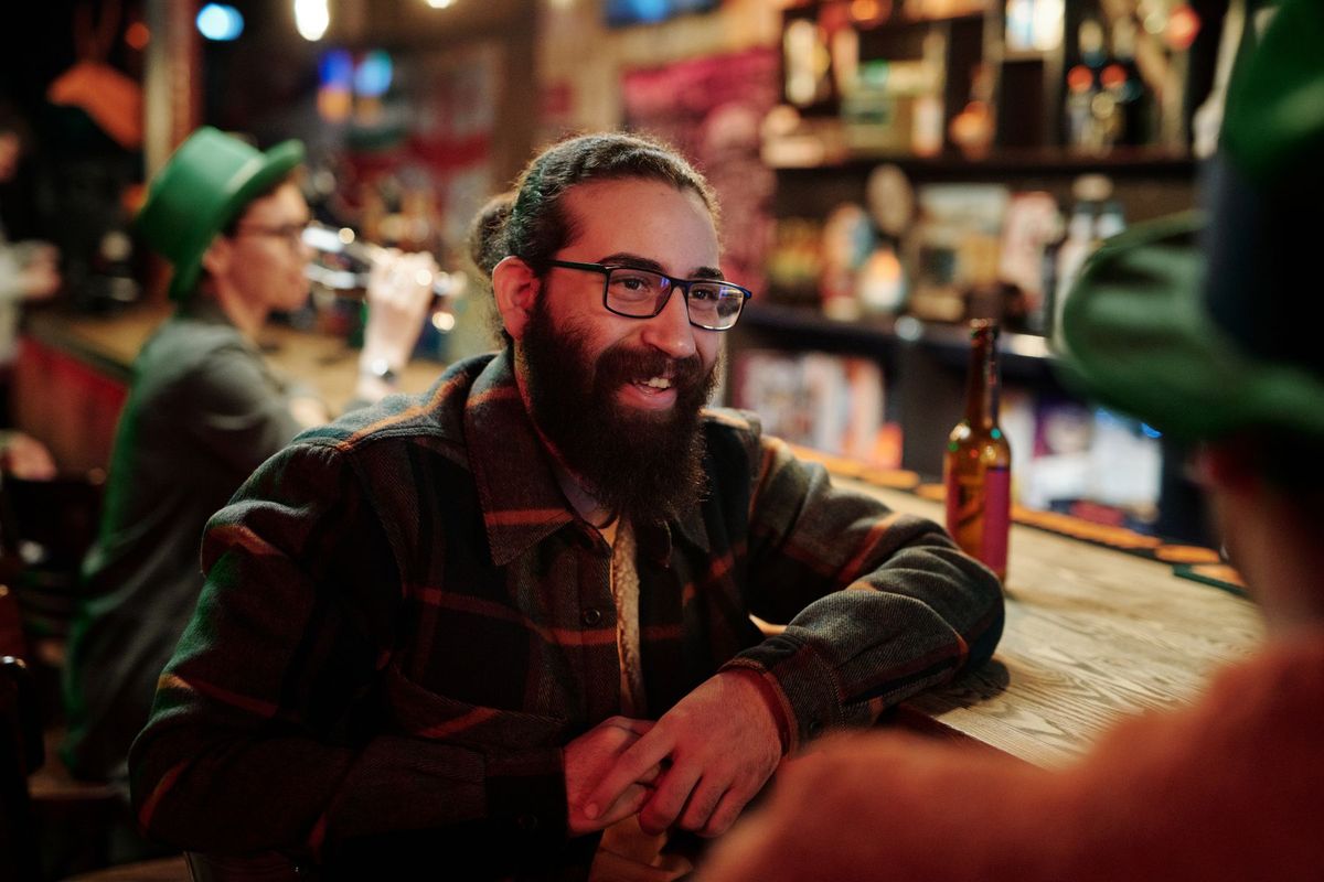 Homme barbu en lunettes discutant avec des clients dans une ambiance chaleureuse de bar rustique