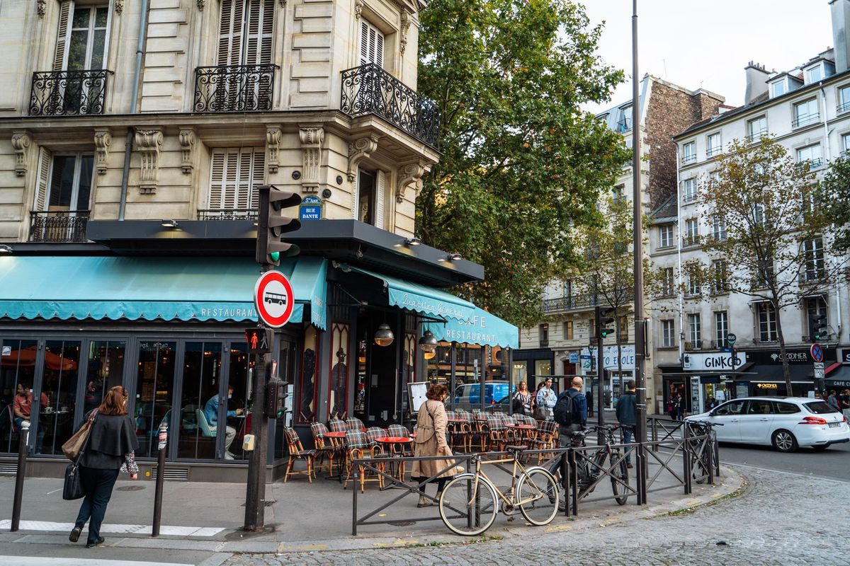 Terrasse de café parisien avec tables en rotin, chaises bistrot et façade haussmannienne sur rue pavée