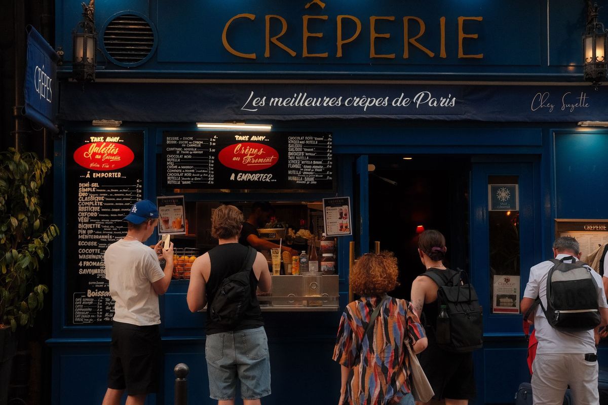 Une file de clients attend devant la façade bleue d'une crêperie parisienne avec ses panneaux de menu visibles