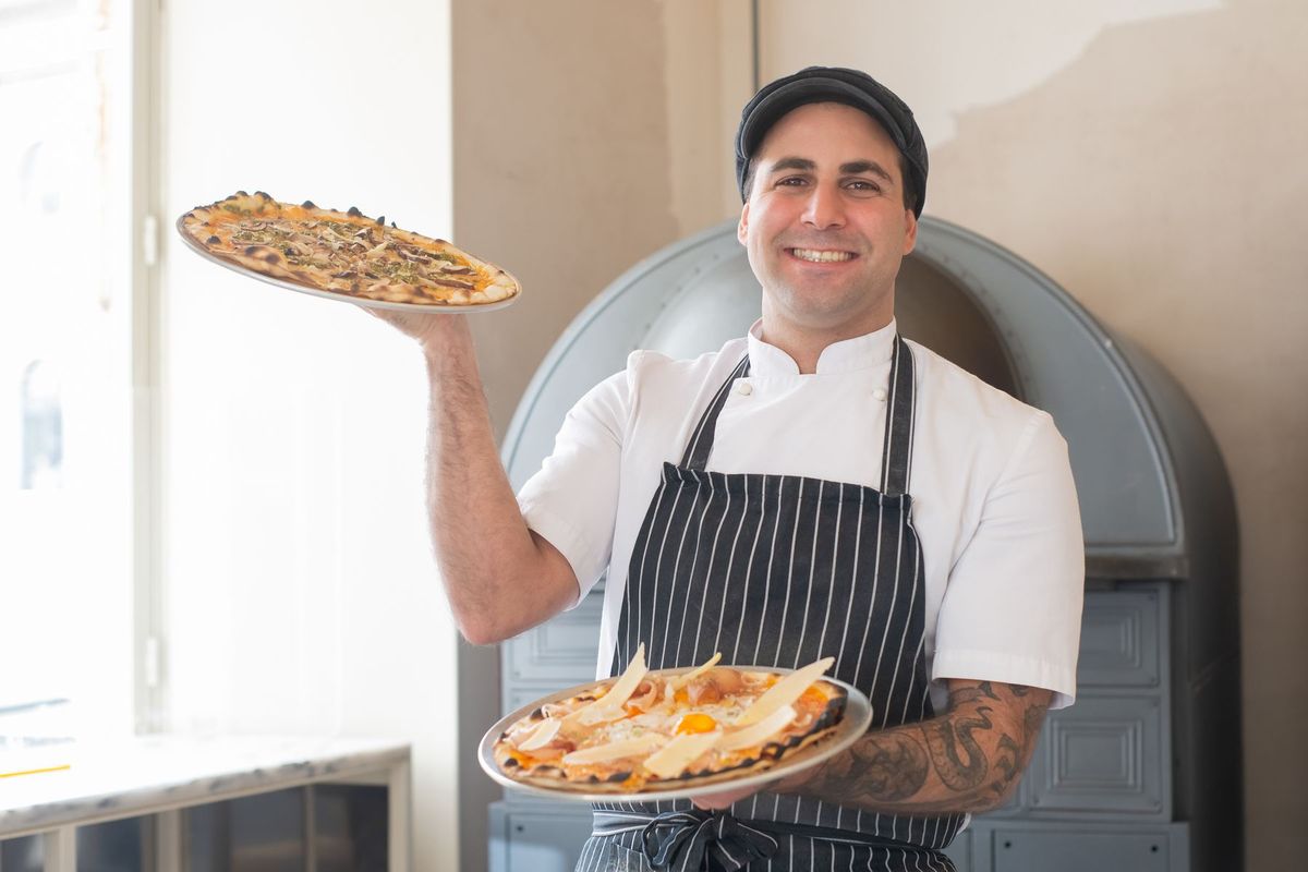 Chef en uniforme blanc présentant des pizzas fraîchement cuites dans son four à bois
