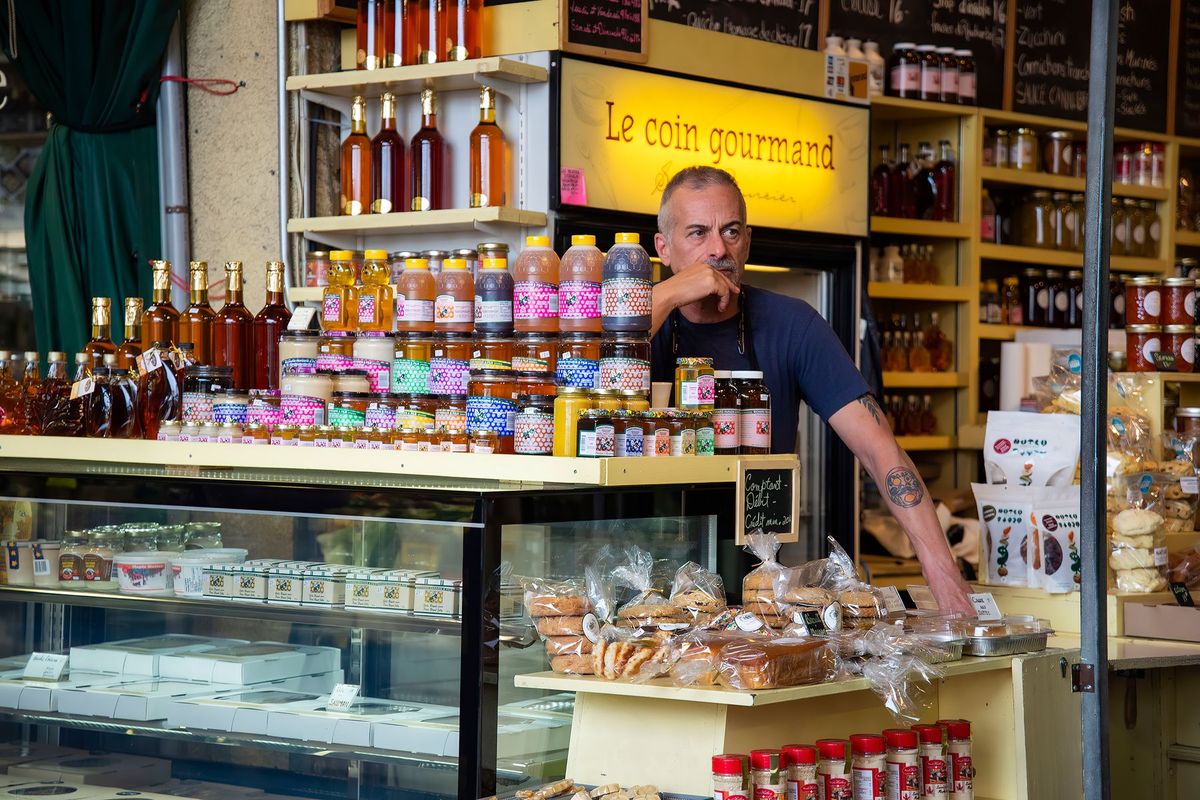 Commerçant derrière son étal de marché présentant fruits, légumes et produits gastronomiques variés