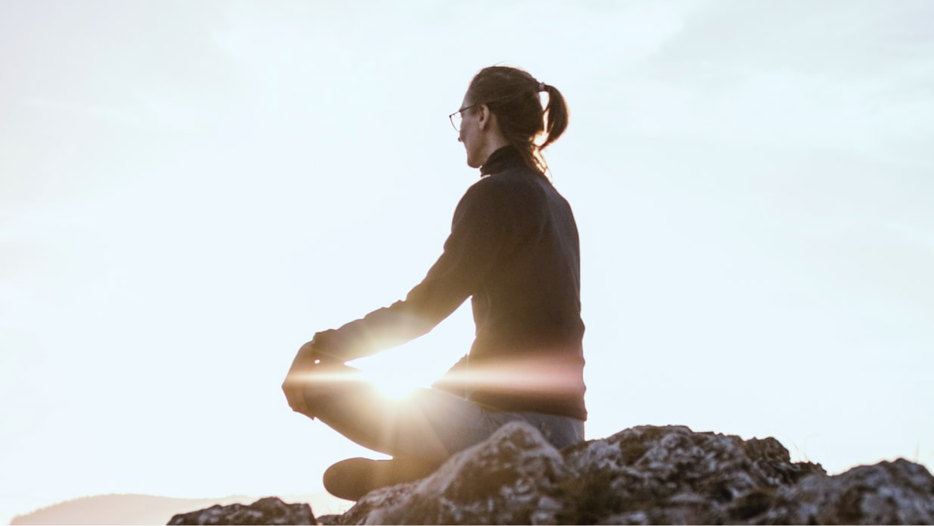 A woman sits mindfully meditating on a rock with sunlight filtering in around her.