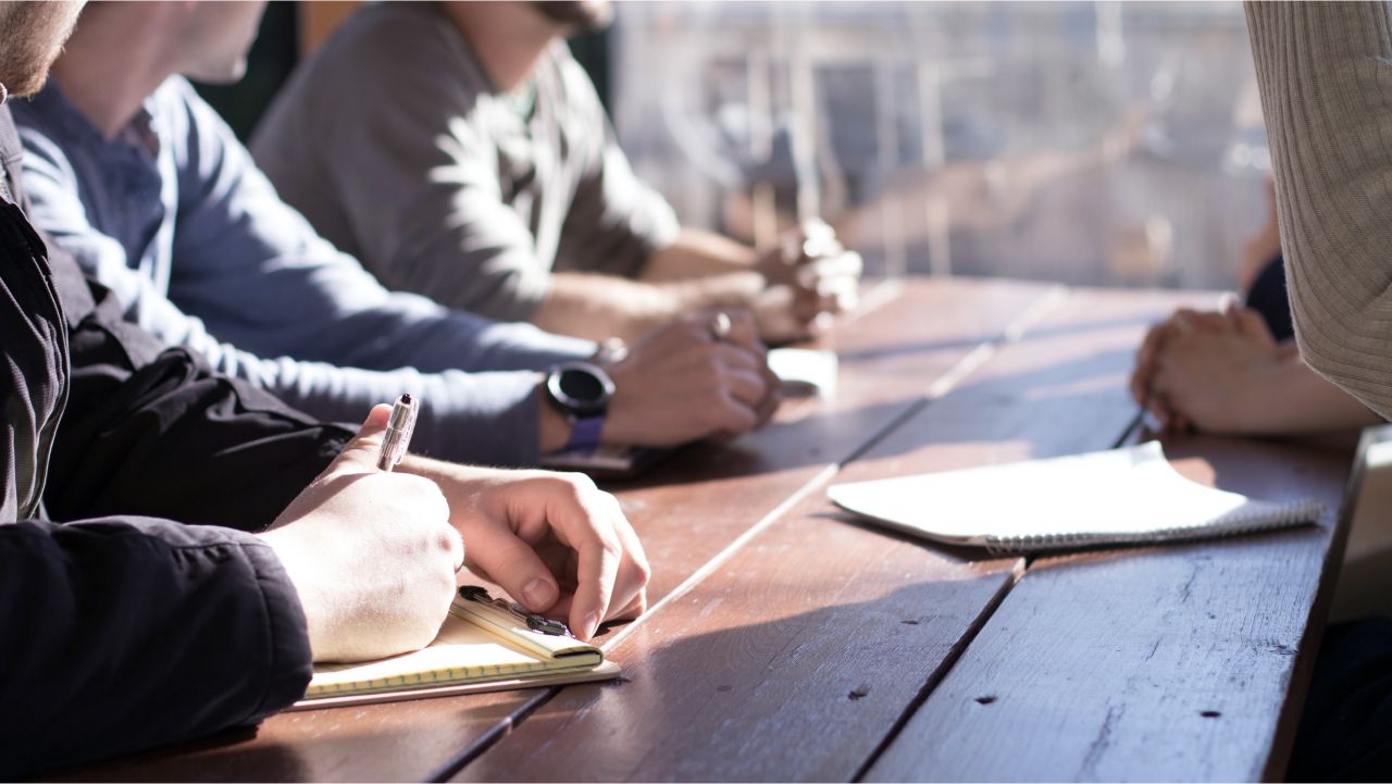 A team gathers around an wooden table, their hands  and some papers on the table as the meet to discuss their next project (and how not to procrastinate on it).