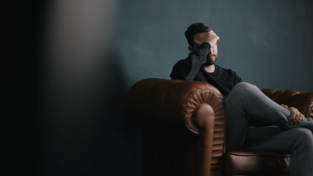 Absenteeism is often the result of poor mental wellbeing. A man sits in a dark room on a sofa, covering his face.