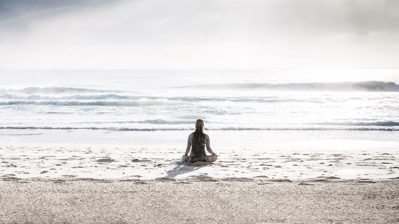 A person with long hair sits on a sandy beach in front of the ocean, meditating and focusing on their breath to relax the body.