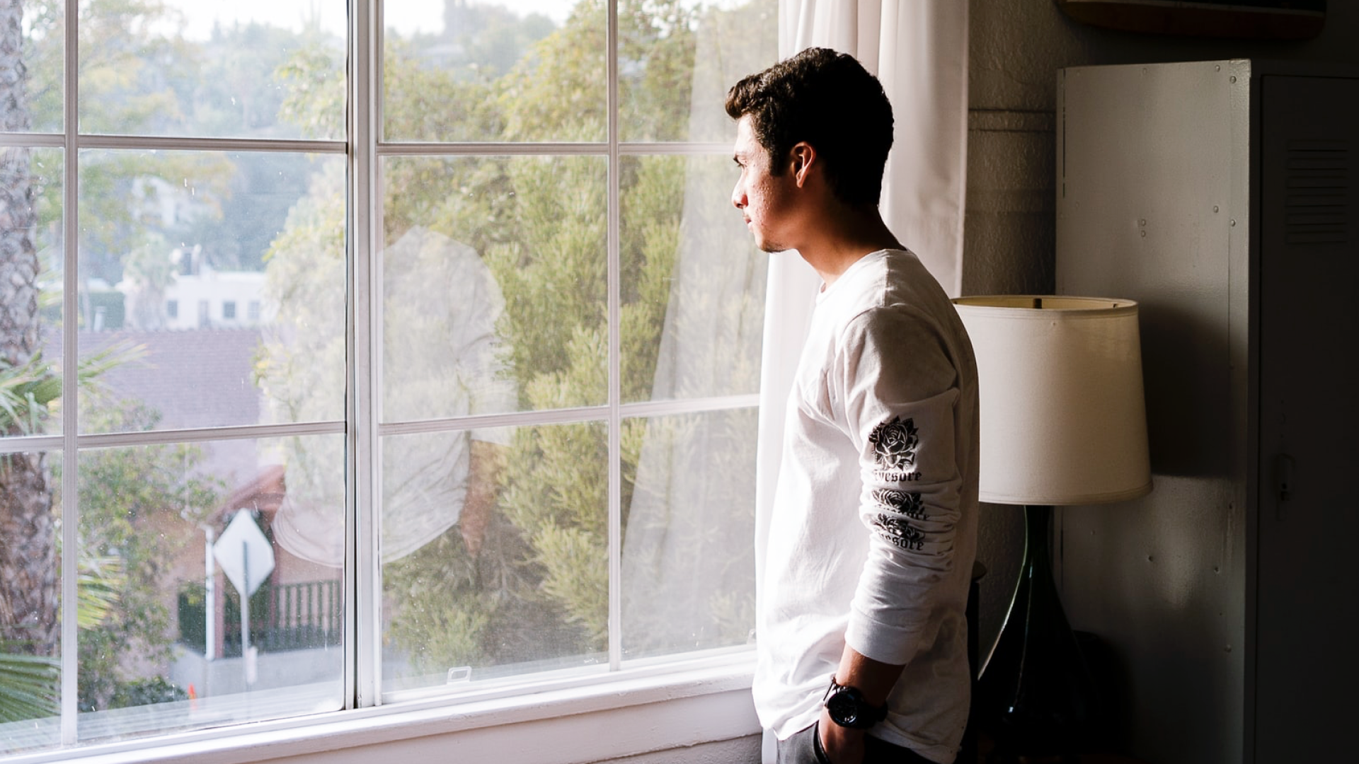 A young person looks out the window at a tranquil, verdant street, moving their focus back to the present moment during the grieving process.