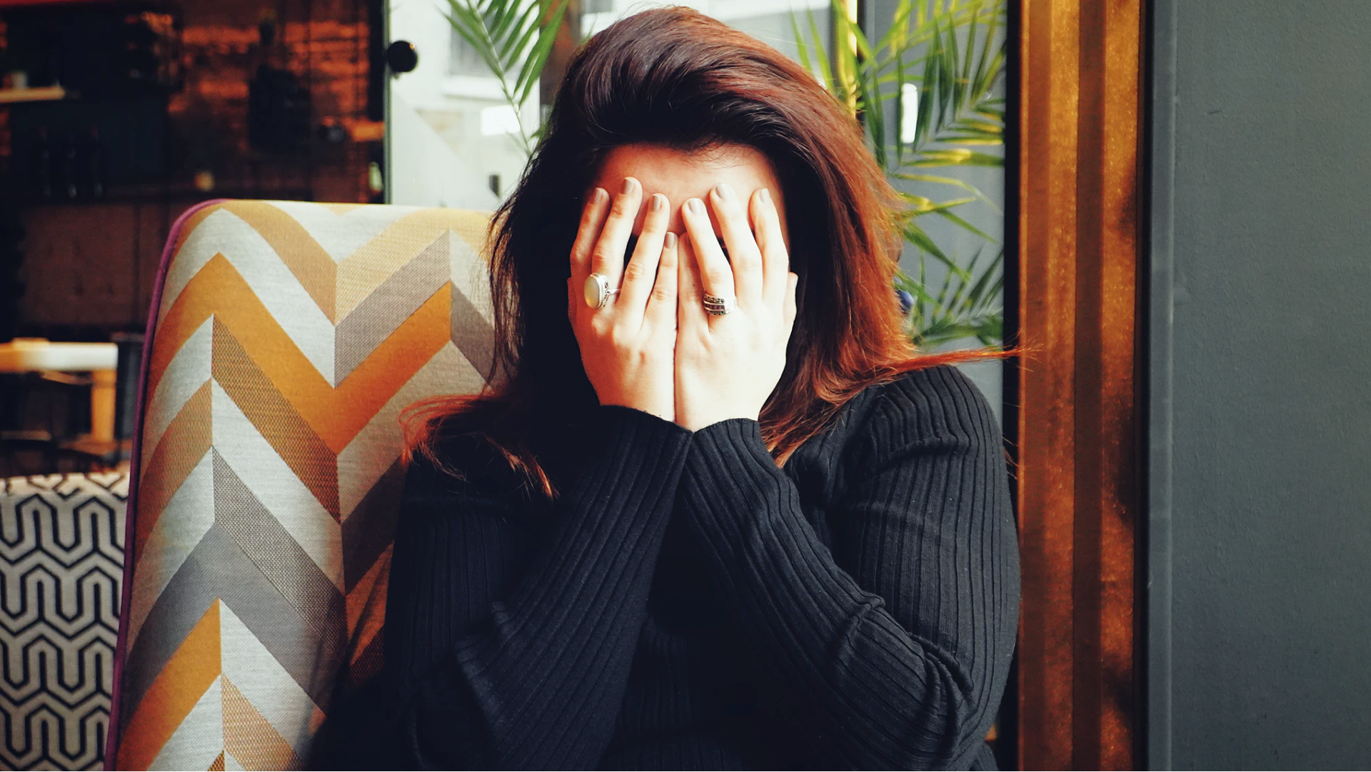 A stressed woman (who's maybe ruminating) covers her eyes with her hands, as she sits in a chair upholstered with chevron fabric.