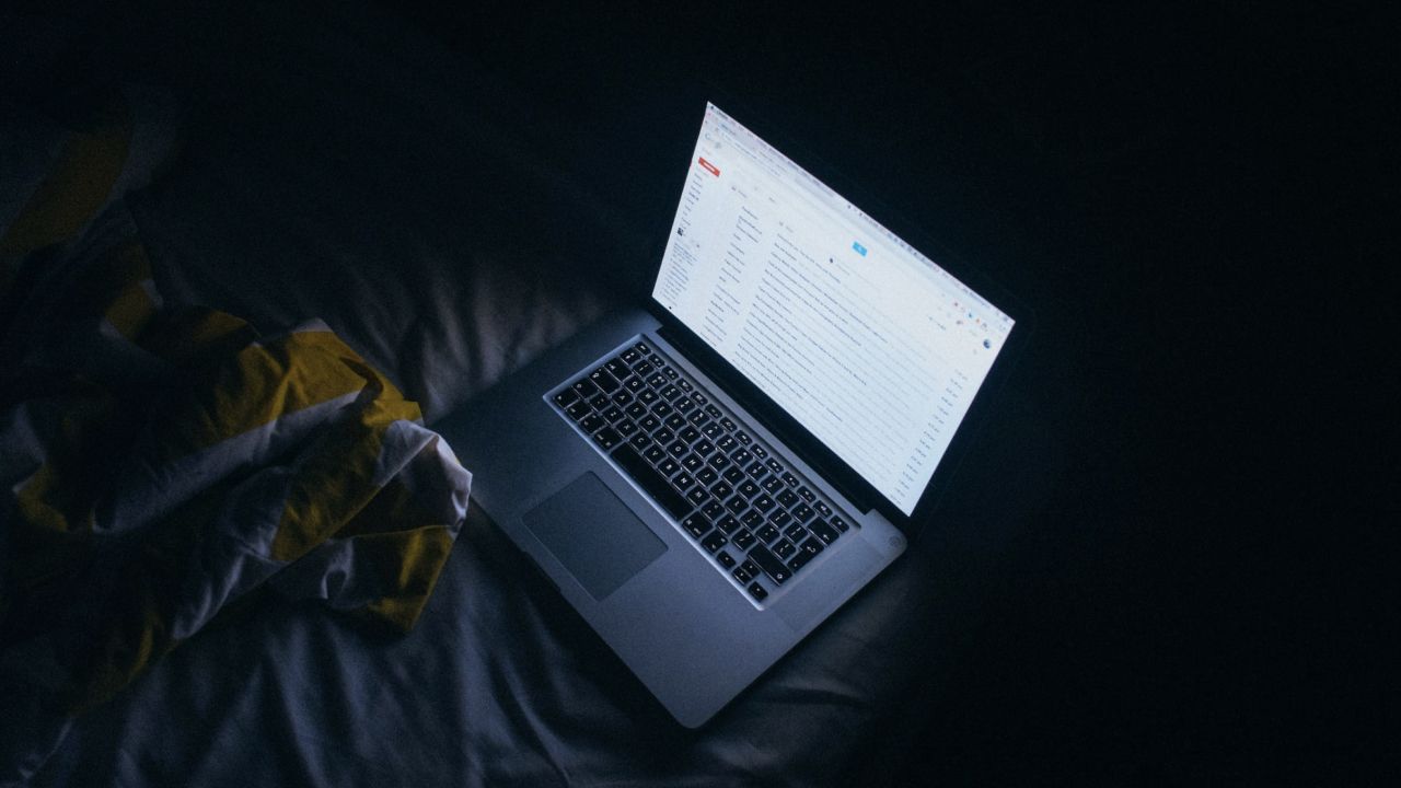 A computer screen sits on a bed next to folded winter coat, illuminating the darkness.