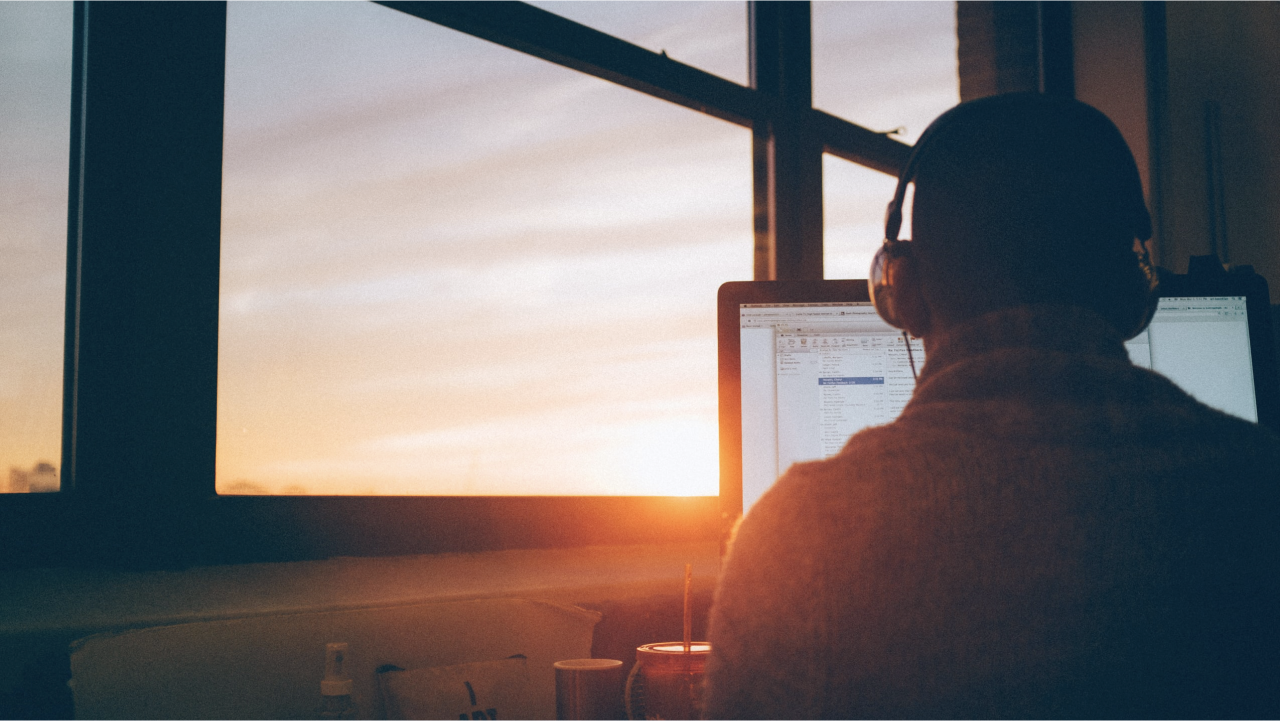 An employee works in front of a screen with headphones on, seemingly oblivious to the sunset beyond him, potentially because of stress.