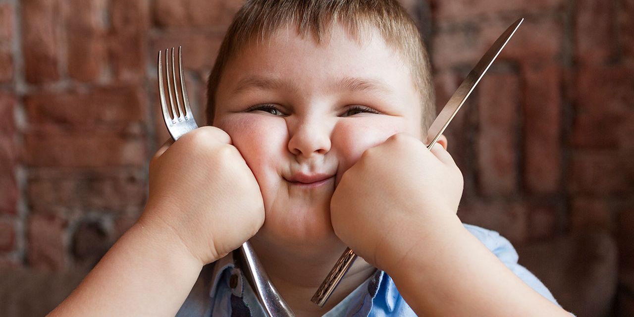 Criança sorridente com excesso de peso segura faca e garfo, ilustrando tema sobre obesidade infantil e alimentação